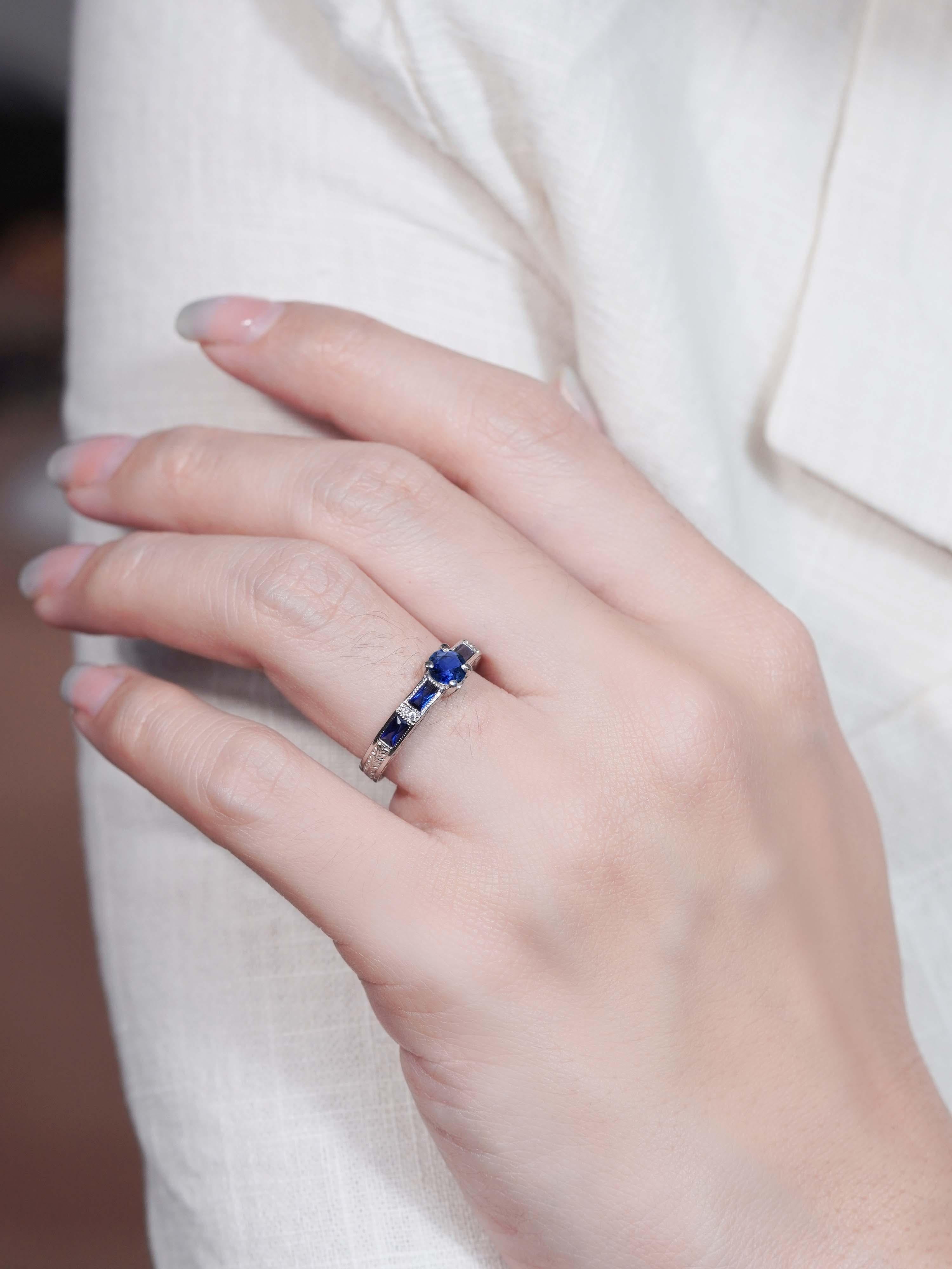 Cette bague de fiançailles solitaire de style Brilliante présente un saphir rond brillant, serti en pointe pour mettre en valeur sa clarté. Les épaules sont ornées de saphirs bleus de taille française, ajoutant une couleur riche et une élégance