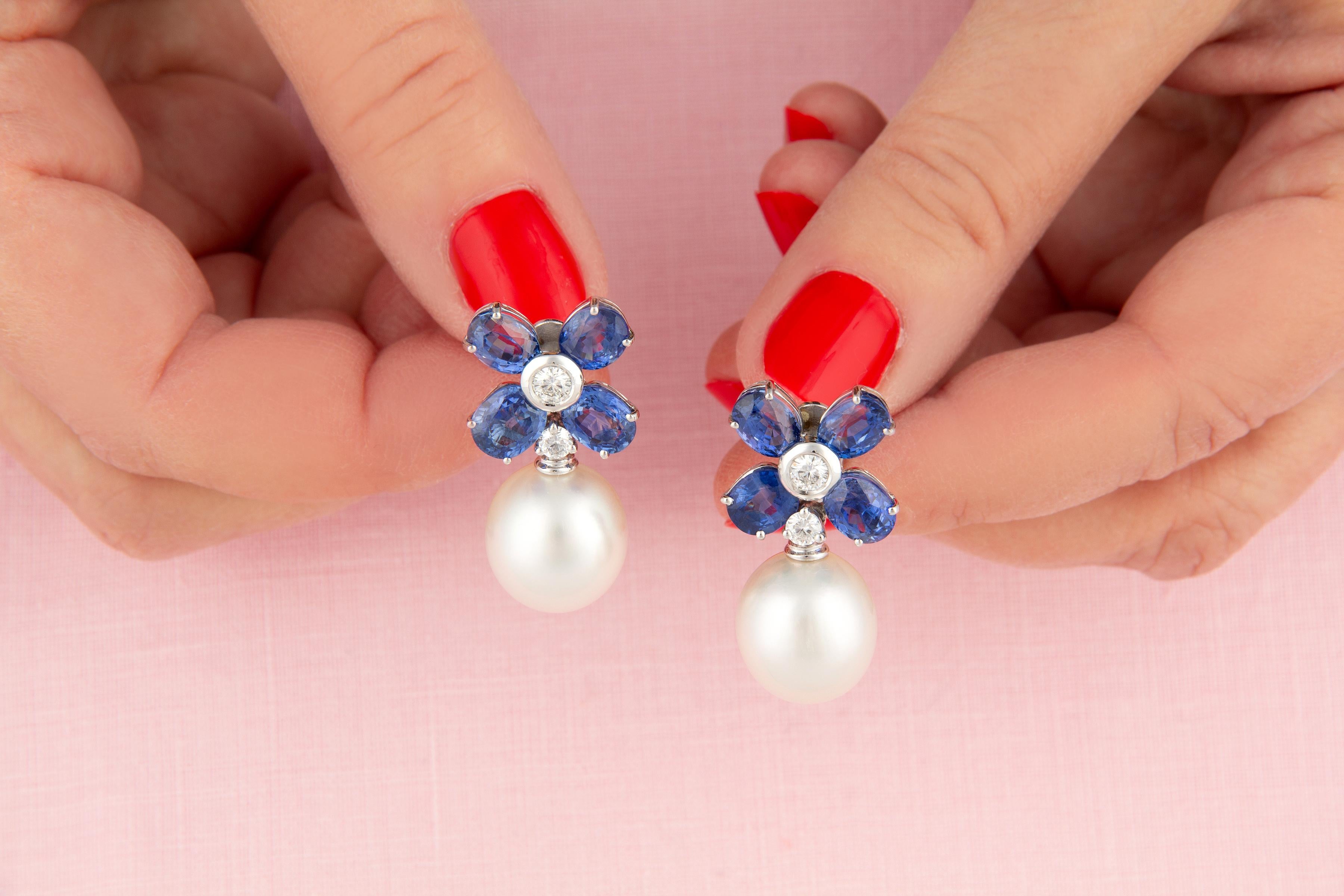 This pair of blue sapphire and pearl earrings features a flower design on the ear with 12.71 carats of oval cut faceted Ceylon sapphires of brilliant blue color. The tops suspend 2 South Sea pearls of 14 x 13mm diameter. The design is complete with