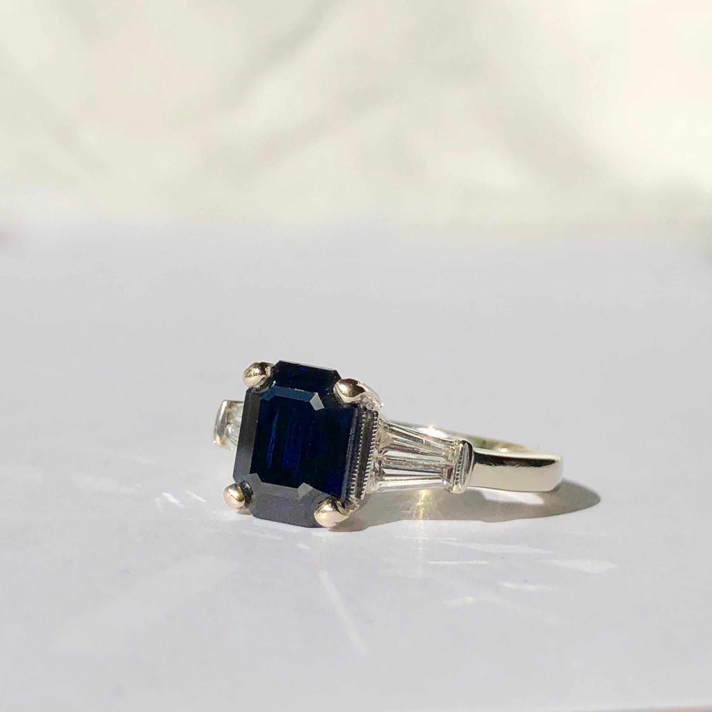 An Emerald Step Cut Midnight Blue Sapphire vintage C1980-90's 

A deep blue hue sapphire set in a classic, elegant style mount fashion from polished 18k white gold with white tapered baguette shoulders

The beautiful white diamonds are and estimated
