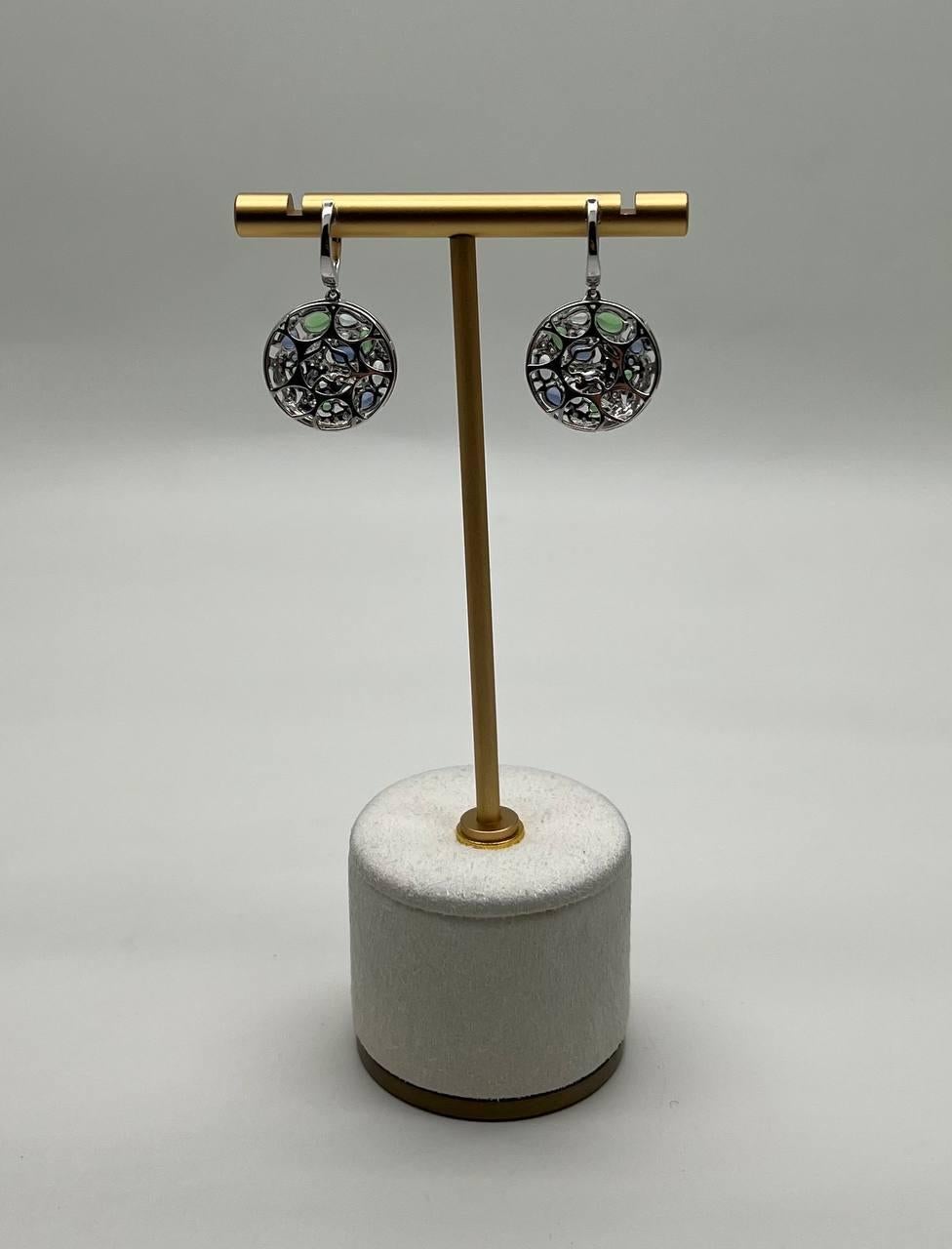 These exquisite earrings feature a circular design adorned with vibrant blue sapphires and green emeralds. The intricate arrangement of stones creates a captivating mosaic effect. The earrings are crafted with precision, showcasing a harmonious