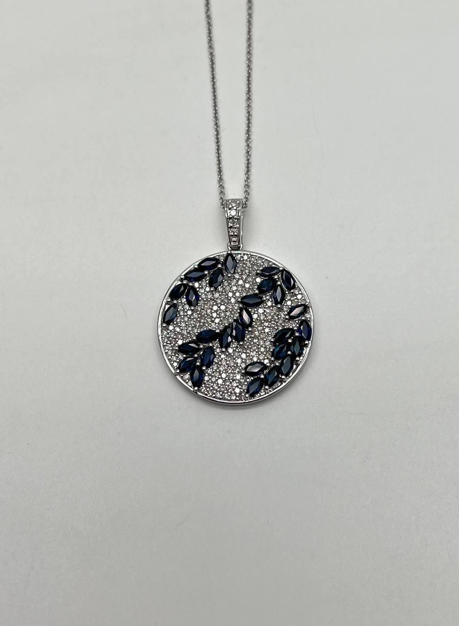 An exquisite pendant necklace. The round pendant features a delicate arrangement of dark blue marquise-shaped sapphires set against a shimmering backdrop of white, sparkling diamonds. The intricate design is framed in a polished metal setting,