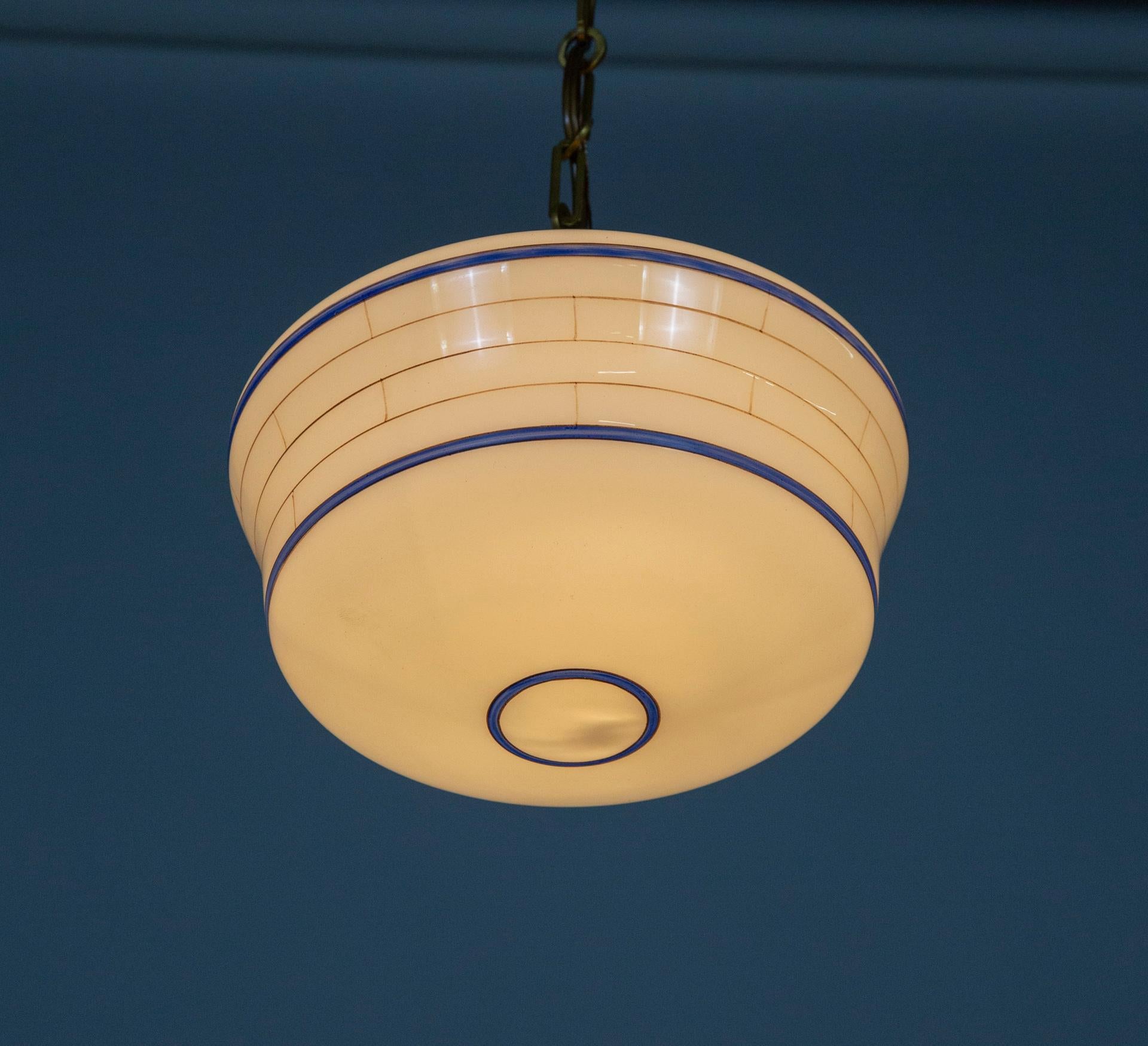 A pair of unique, milk glass pendant lights are hand painted with thin, blue stripes circling the form, and very subtle, hairline, gold brick-form stripes on the sides. These highly pleasing, antique glass pieces are hung with new brass shade