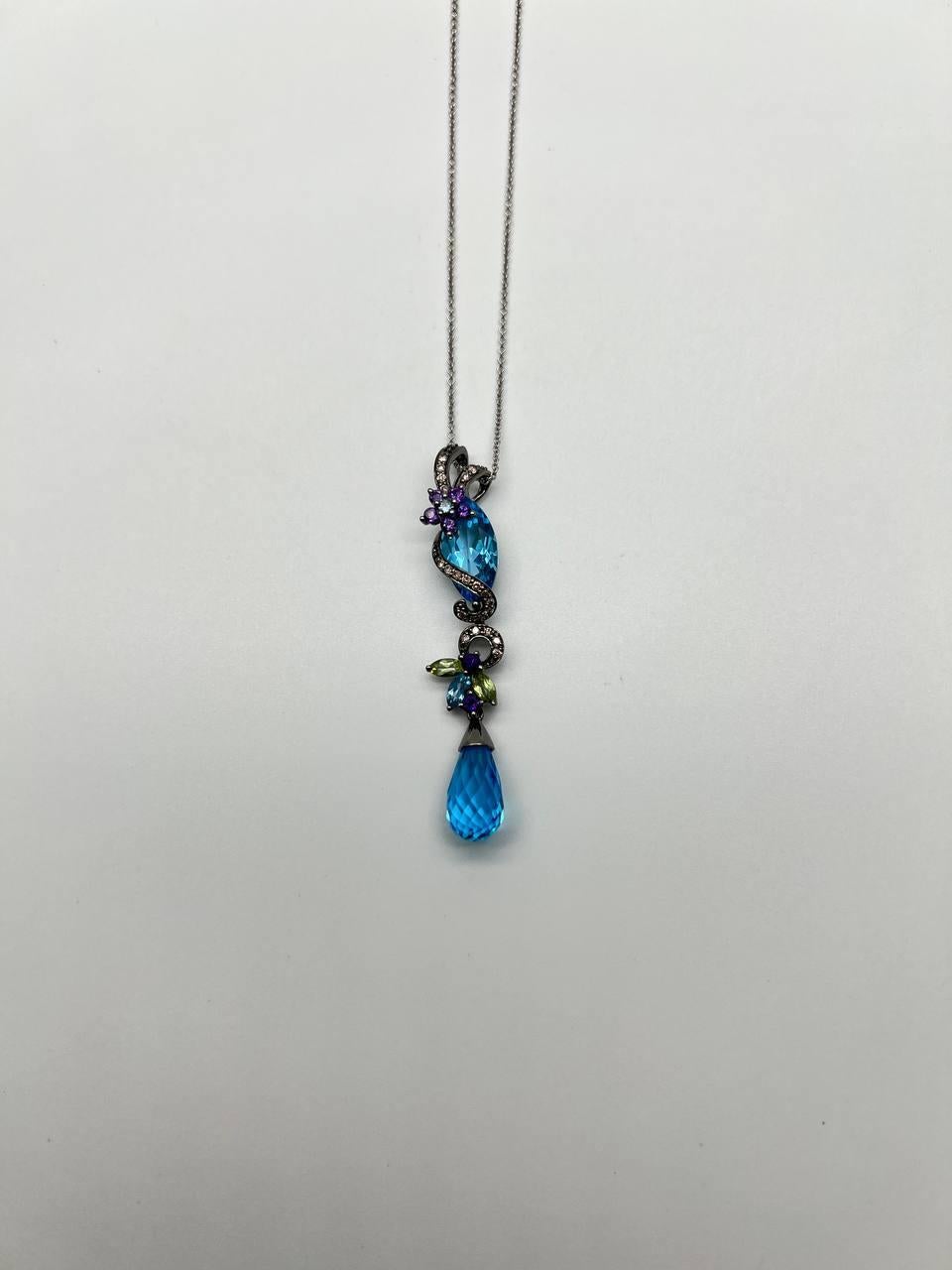 Este elegante collar colgante, presenta un llamativo diseño con un vibrante topacio azul en forma de lágrima, expertamente facetado para captar la luz. Arriba, una serie de gemas más pequeñas en amatista morada y peridotos verdes están dispuestas de