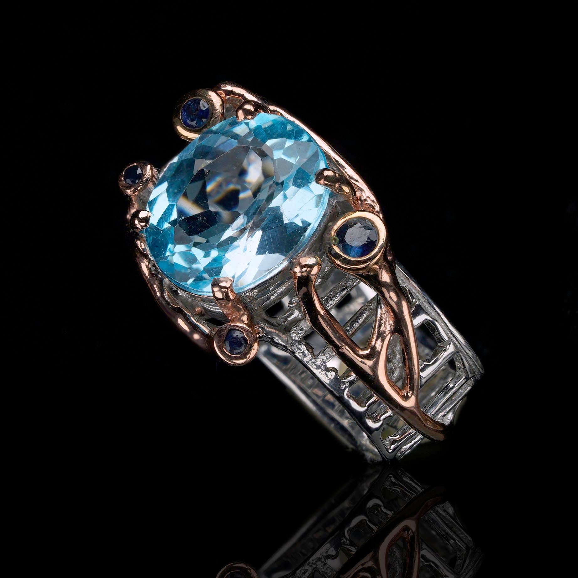 This stunningly unique artisan-crafted, eminently giftable ring features a brilliant blue Topaz at its center cradled by a sculptural setting of sterling silver and rose gold accented with blue Sapphires.

The radiant oval-cut Topaz,