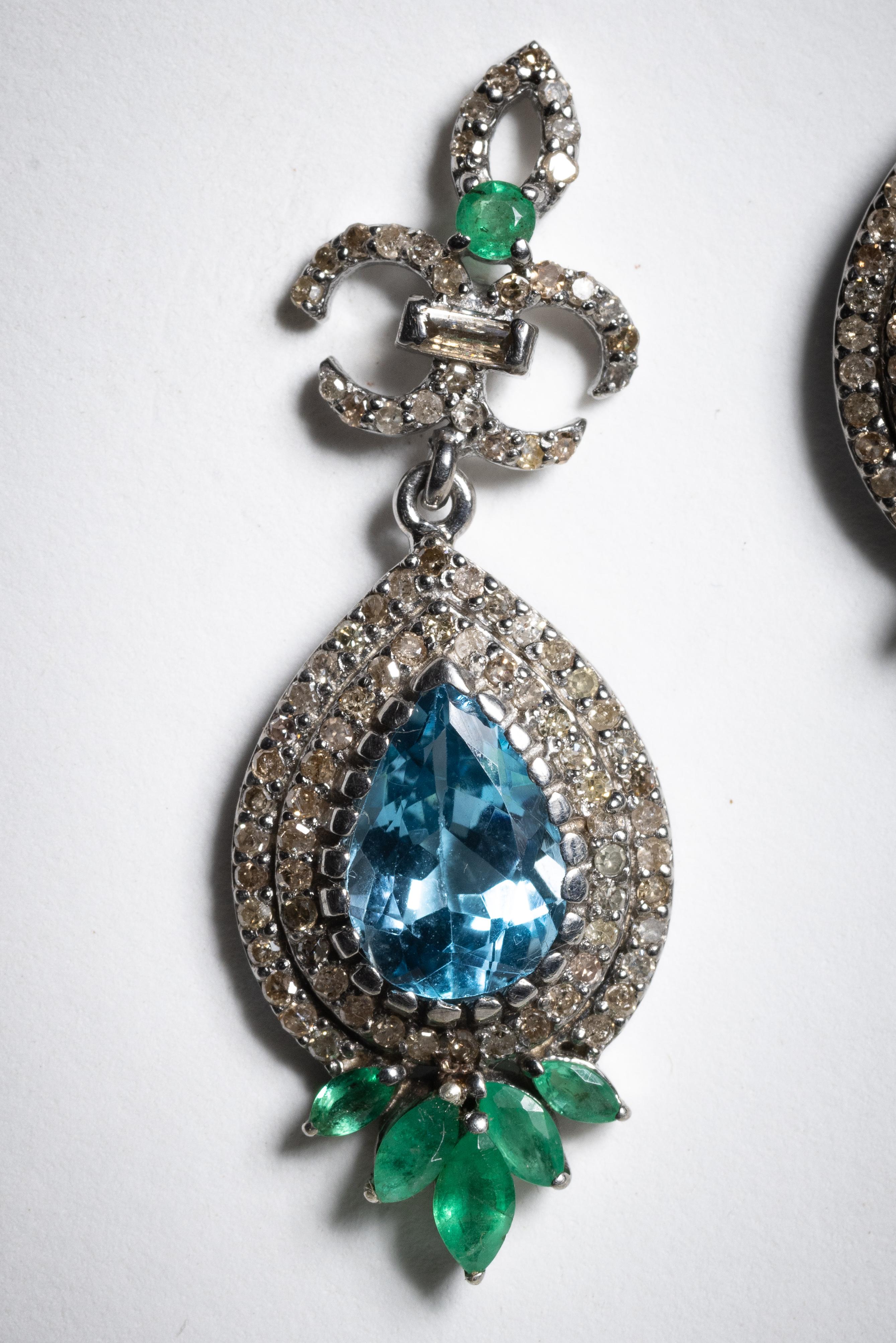 A stunning pair of large, facetd pear-shaped blue topaz gemstones in a bezel setting bordered with a double row of round, brilliant cut diamonds.  At the bottom each earring has a graduated cluster of 5 marquise cut emeralds.  Set in sterling silver