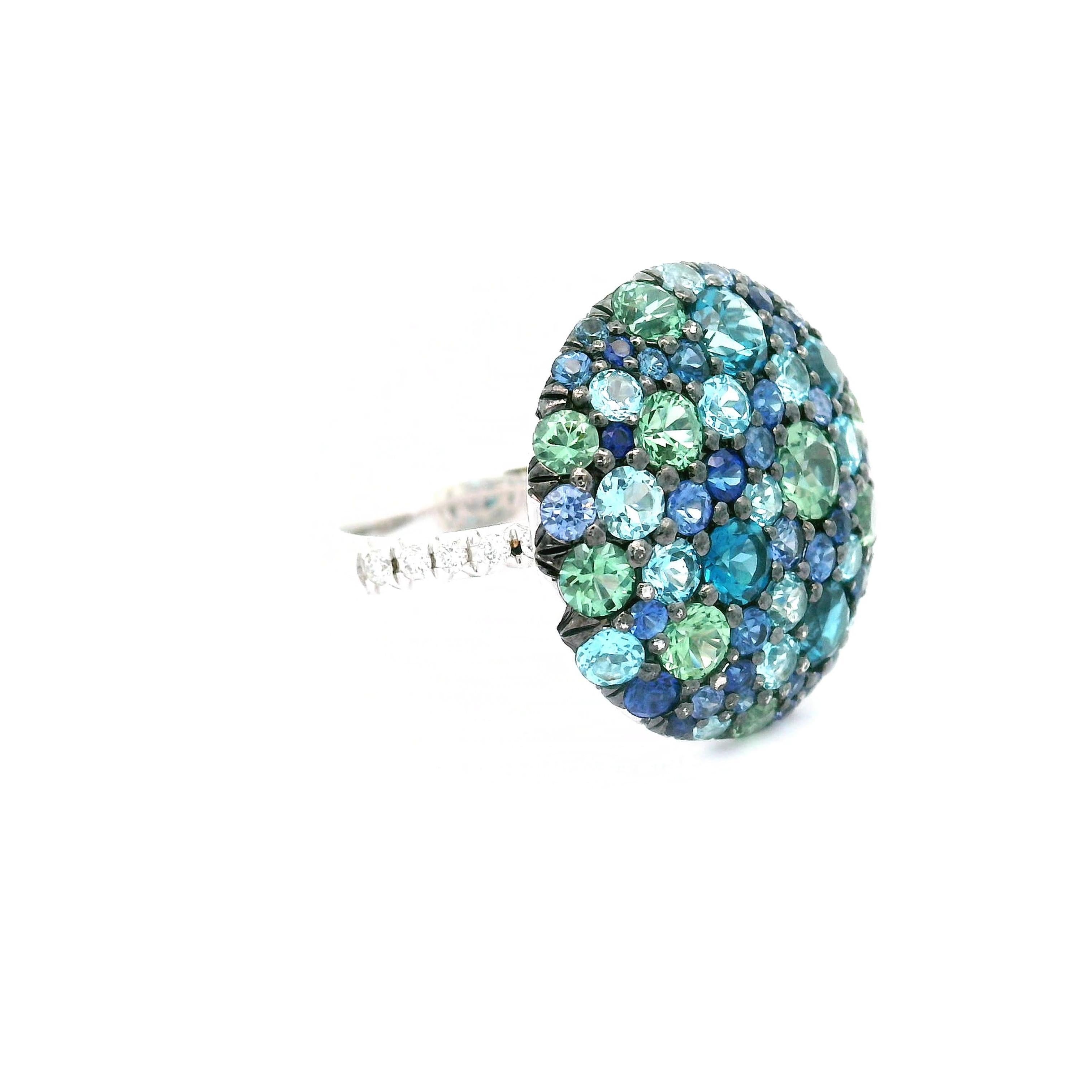 A stunning contemporary ring featuring a 1.83 carat round blue topaz as its centerpiece. The topaz exhibits a vibrant blue hue, surrounded by 1.02 carats of round green sapphires, adding a refreshing contrast. Accentuating the design are sparkling