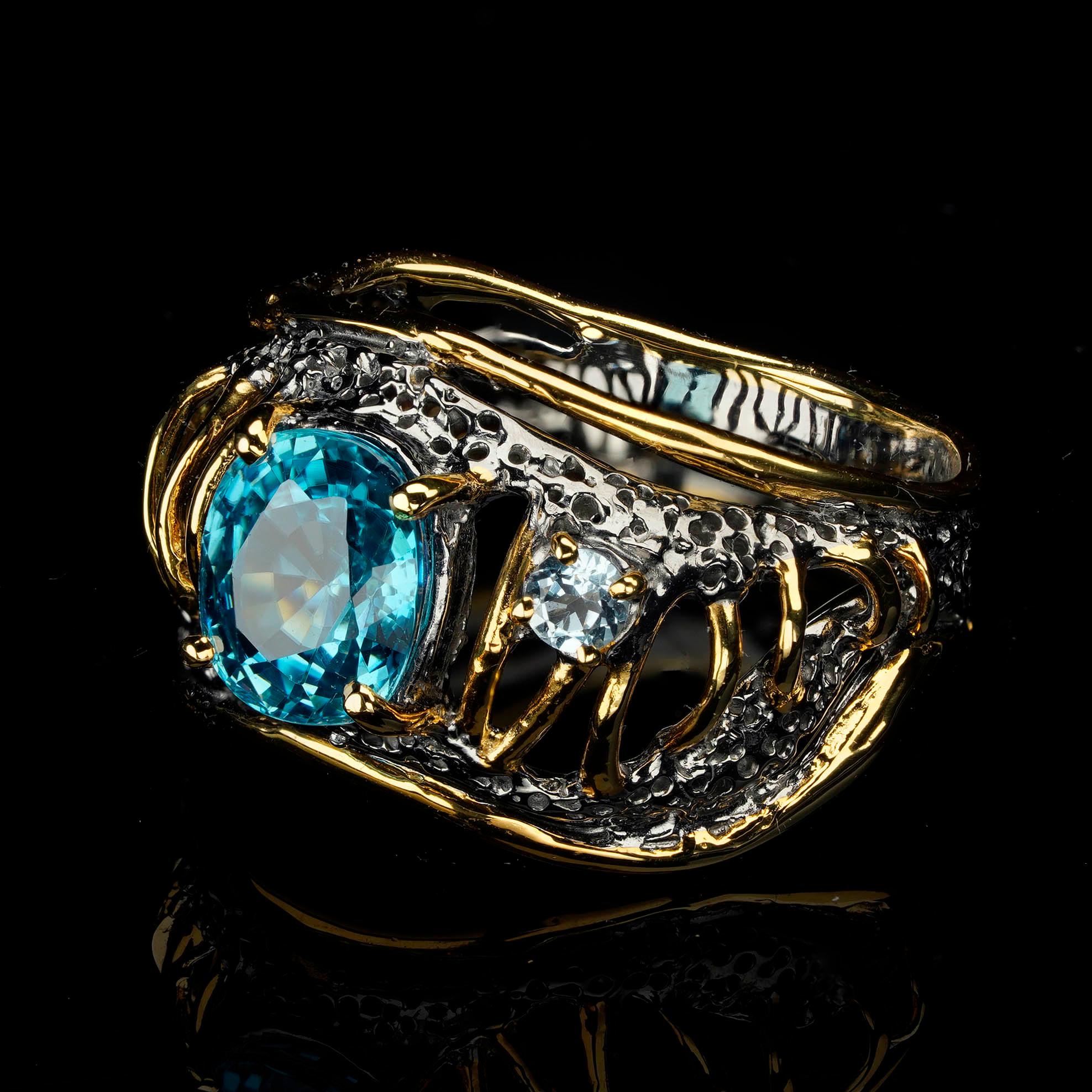 Dieser einzigartige, skulpturale Ring aus Sterlingsilber trägt in seiner Mitte einen blauen Topas im Ovalschliff von mittlerer Größe und mit lebhafter Pigmentierung. Ein funkelnder Moissanit, umrahmt von goldenen Vermeil-Details, die komplizierte