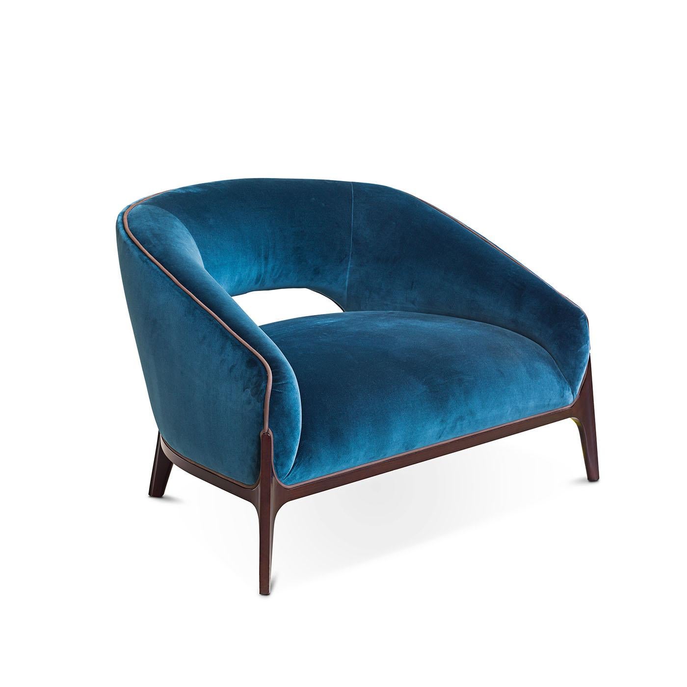This blue armchair is made of velvet upholstery and complemented by a sturdy wooden frame. The leather piping detail along the backrest adds a sophisticated touch, creating a harmonious blend of comfort and elegance.