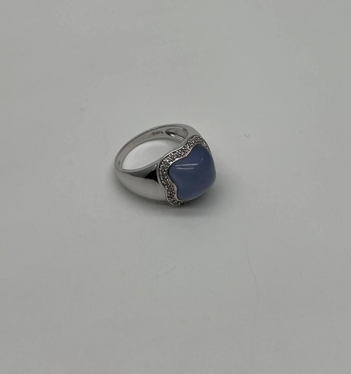 This exquisite piece features a polished white gold band. The centerpiece is a square cabochon stone with a soft blue violet hue, encased in a delicate halo of small, sparkling stones. The setting enhances the stone's gentle color and smooth
