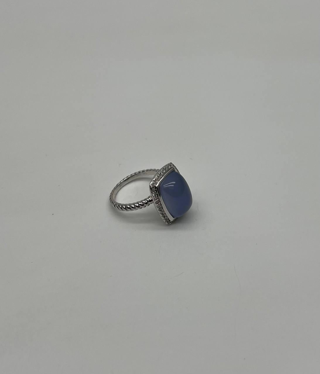 A study in elegance, simplicity, and subtle shine.
This striking ring features a smooth, rectangular blue chalcedony, framed by a delicate halo of sparkling diamonds. The stone's soft hue is beautifully set off by the intricate detailing of the