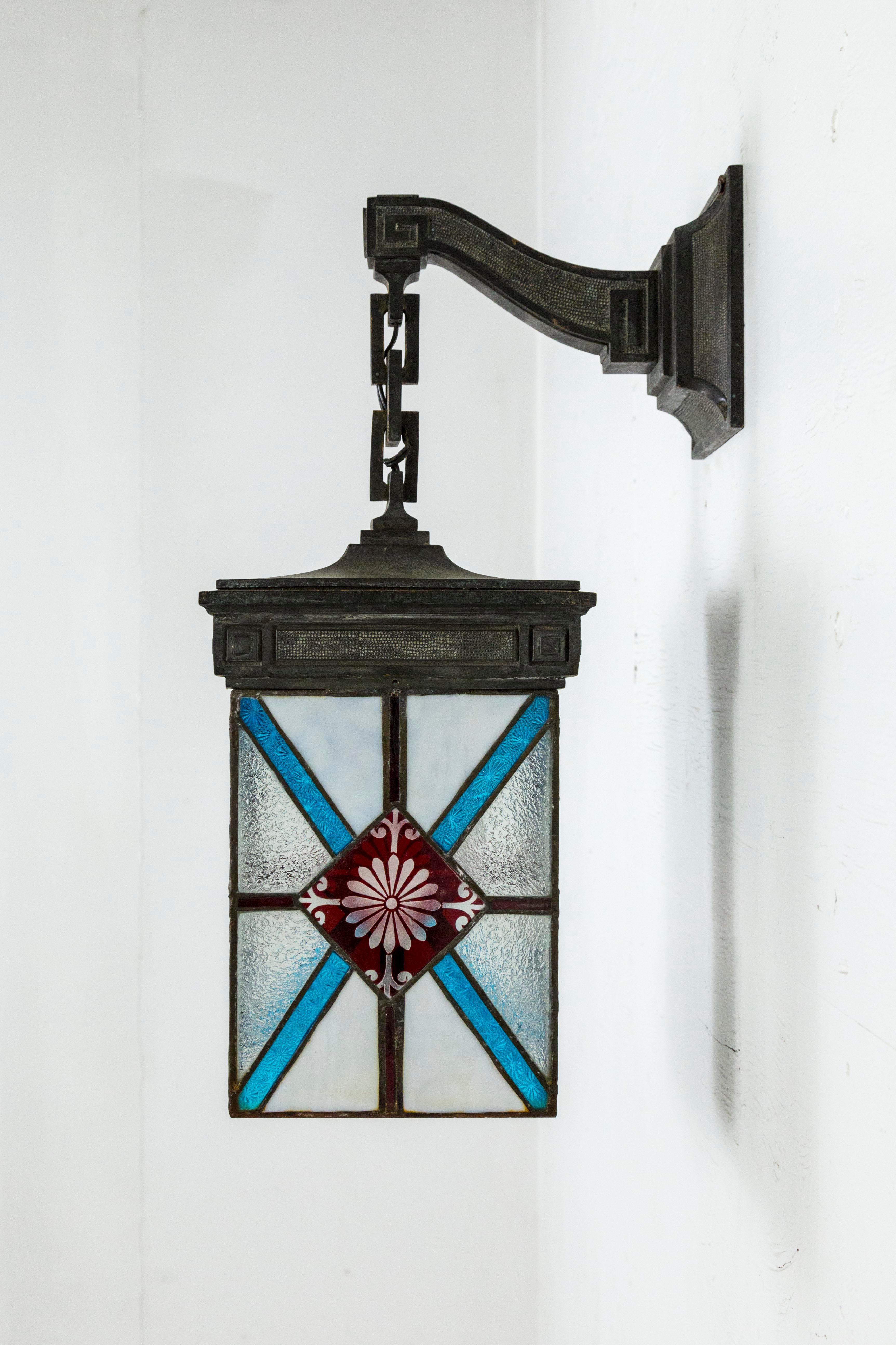 A four-sided, wall hanging, bronze lantern with leaded stained glass in white, blue, and deep red, with textured and cut glass details, diamond and flower designs. The green/gray patina hardware has neoclassical, key elements, and rectangle shaped