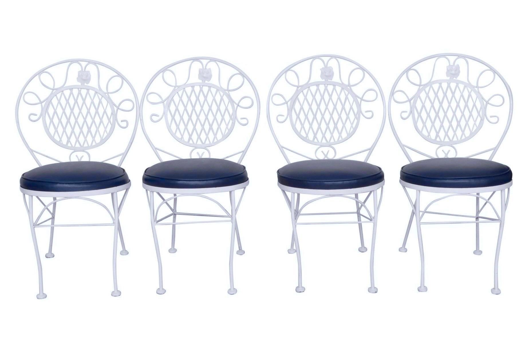 A traditional wrought metal five piece patio dining set in white. The round table is topped with textured glass, set above a scrolled apron finished with floral details. Curved legs are supported with an arched x-stretcher. Four chairs with round