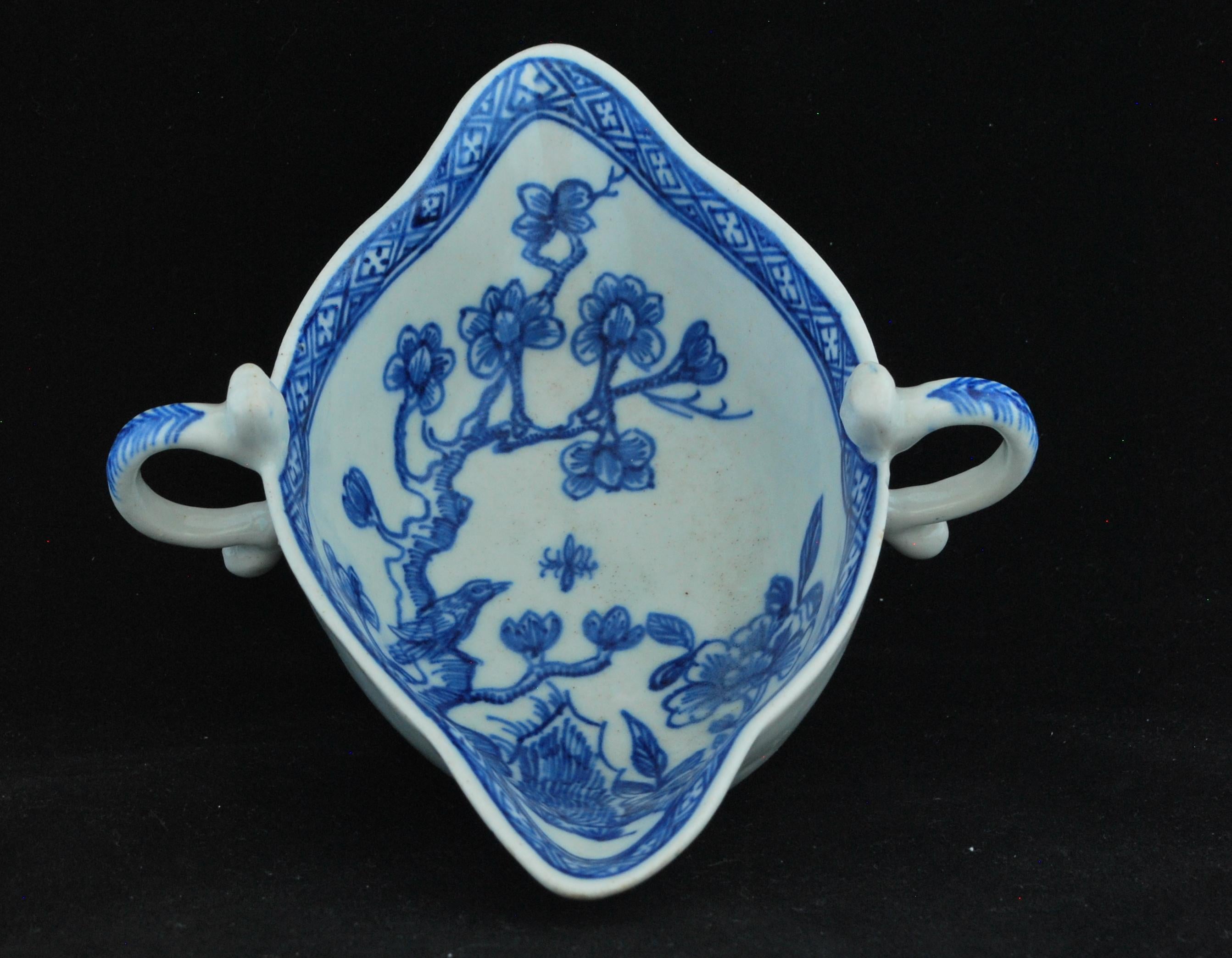 Of silver form; oval, double lipped, and with scroll handle with stylized mask to each side. The interior painted in blue underglaze with a songbird on a branch, a peony and an inner rim diaper border. The exterior of each lip painted with a leafy