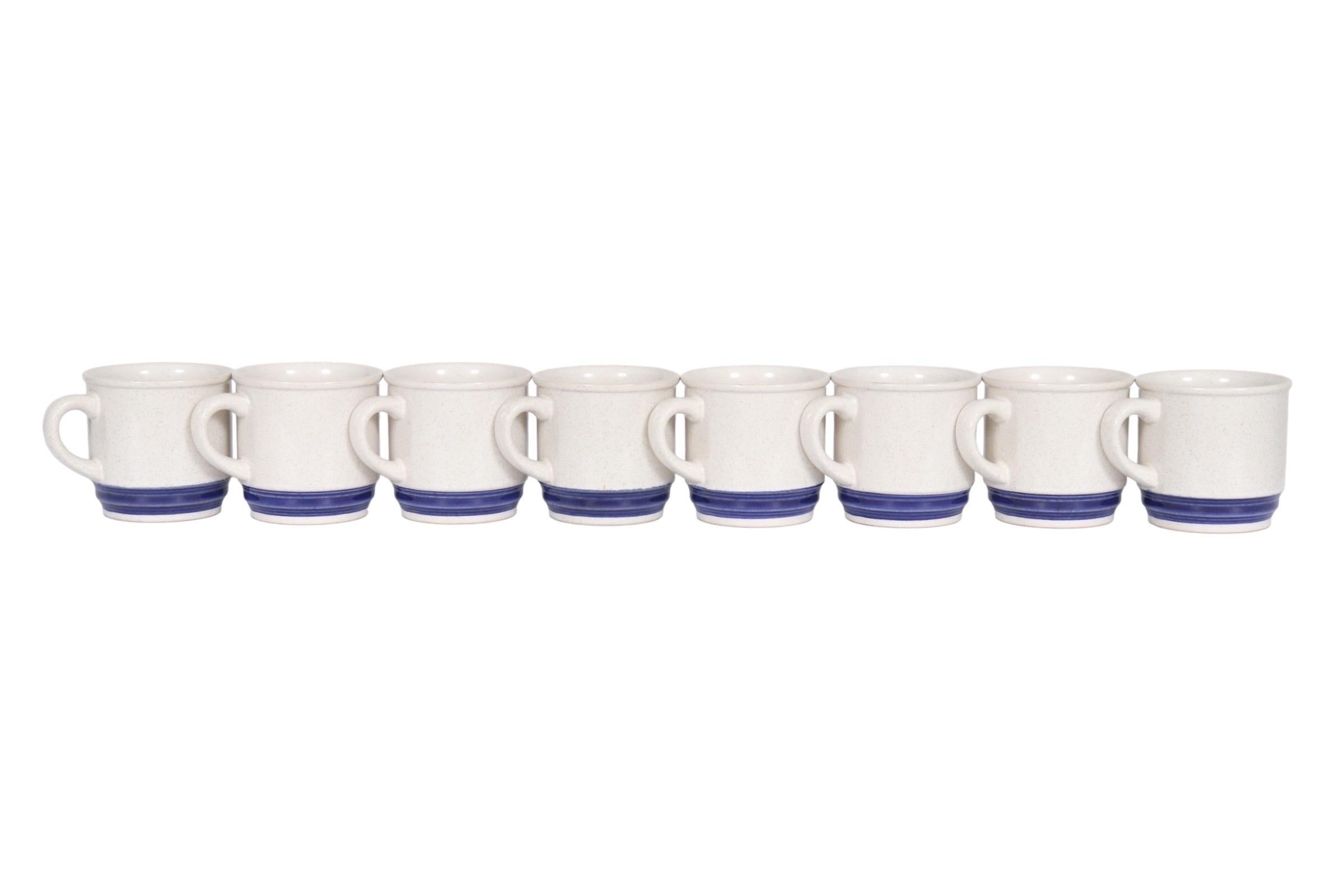 A set of eight stoneware coffee mugs in blue and white. Speckled white stoneware with rounded handles are pressed at the base with a reeded band of cobalt blue. Marked underneath “Stoneware Japan”. Dimensions per mug.