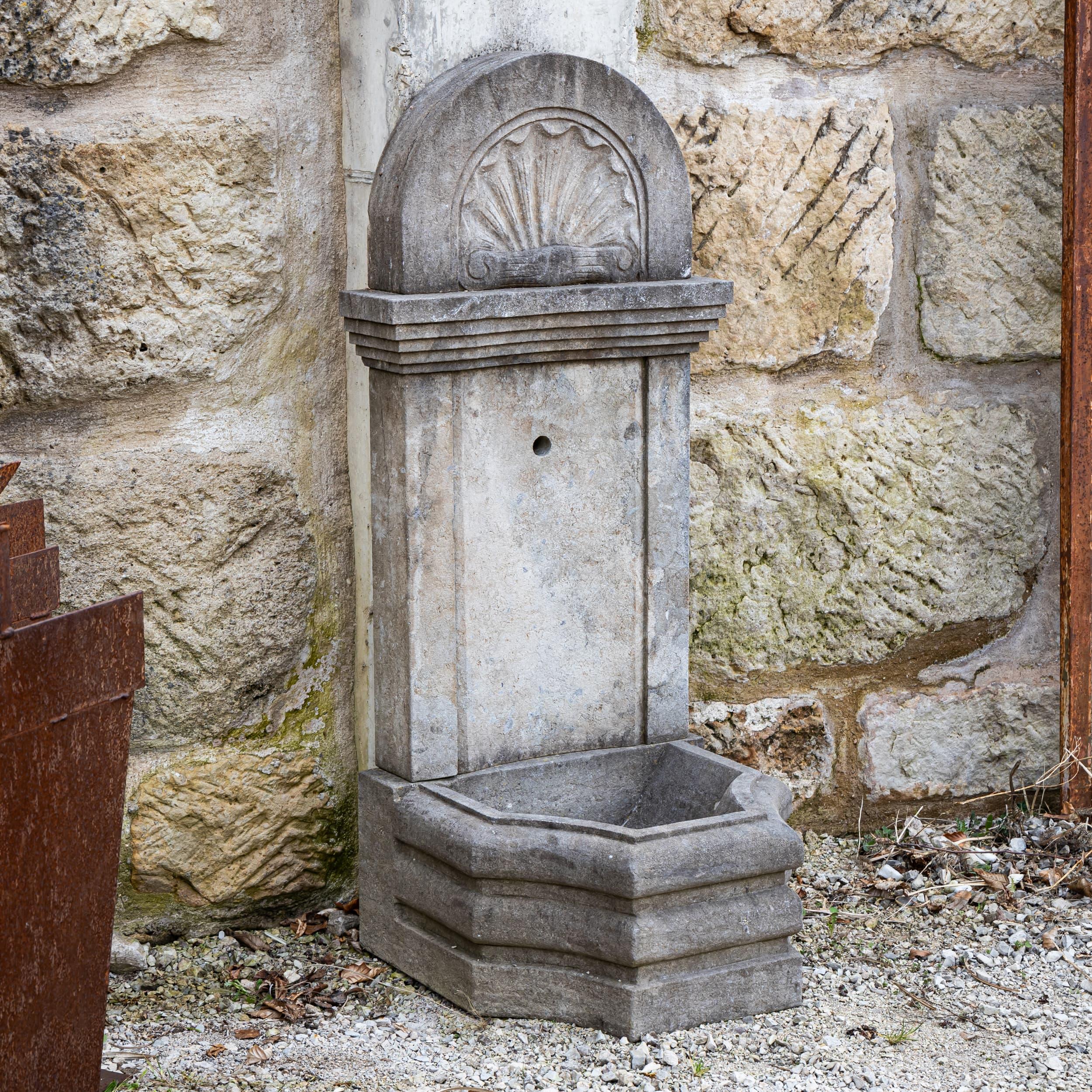 Wall fountain made of hand-chiseled, antique-patinated bluestone with a compact hexagonal basin. The tall back panel with shell ornamentation and a distinctive cornice provides a classic design with high stability.