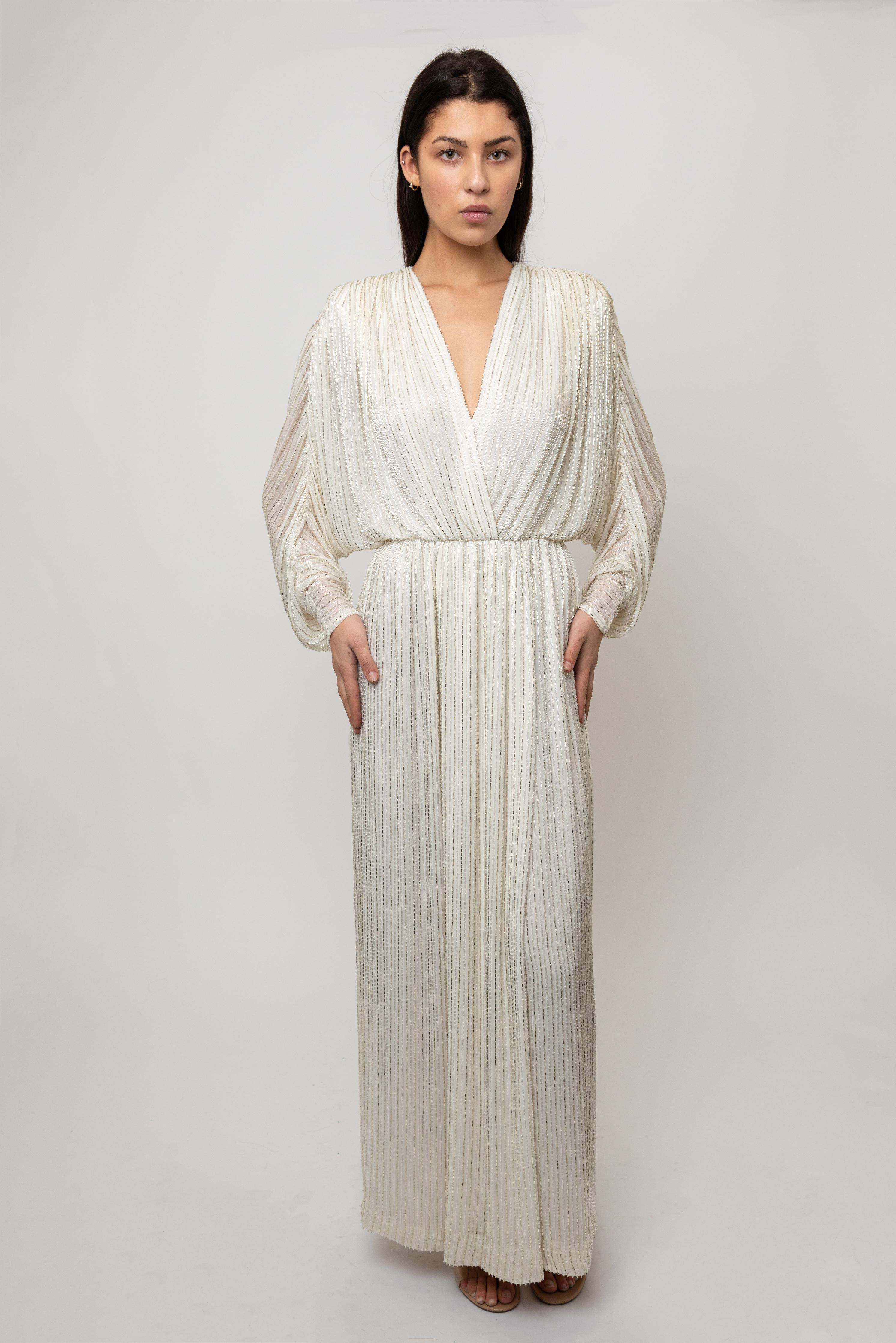 This exquisite Bob Mackie creation epitomizes 1970s glamour with its pristine white silhouette and intricate beadwork. The elegant V-neckline and flowing long sleeves offer timeless sophistication, while the fully beaded construction catches light