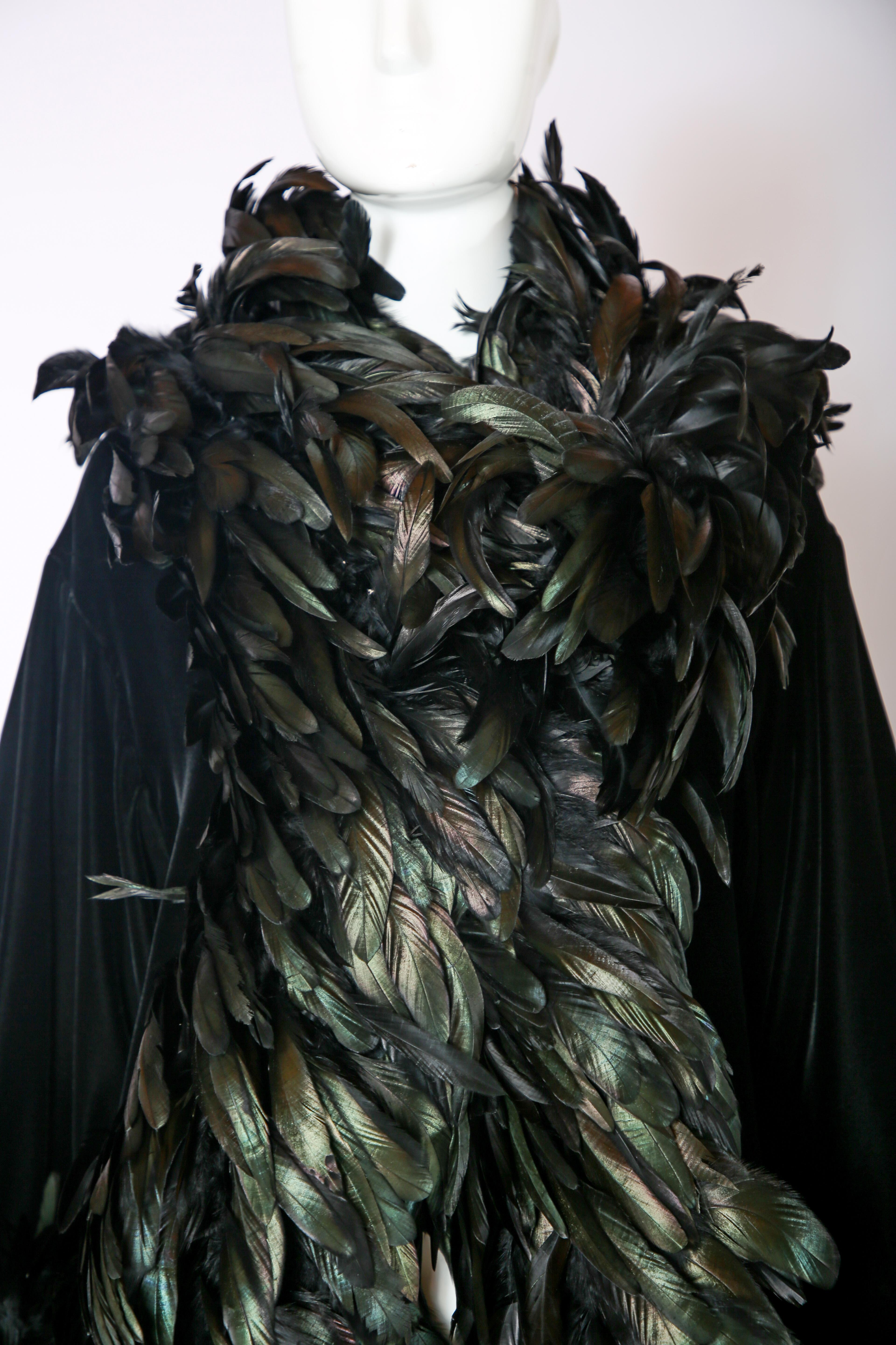 Bob Mackie black velvet coat with black feather trim w/an undertone of green iridescence. There is a hidden hook closure at center front. In very good condition with some scattered and missing feathers. Size 8.

Sleeves: 19” to end of feathers
Bust: