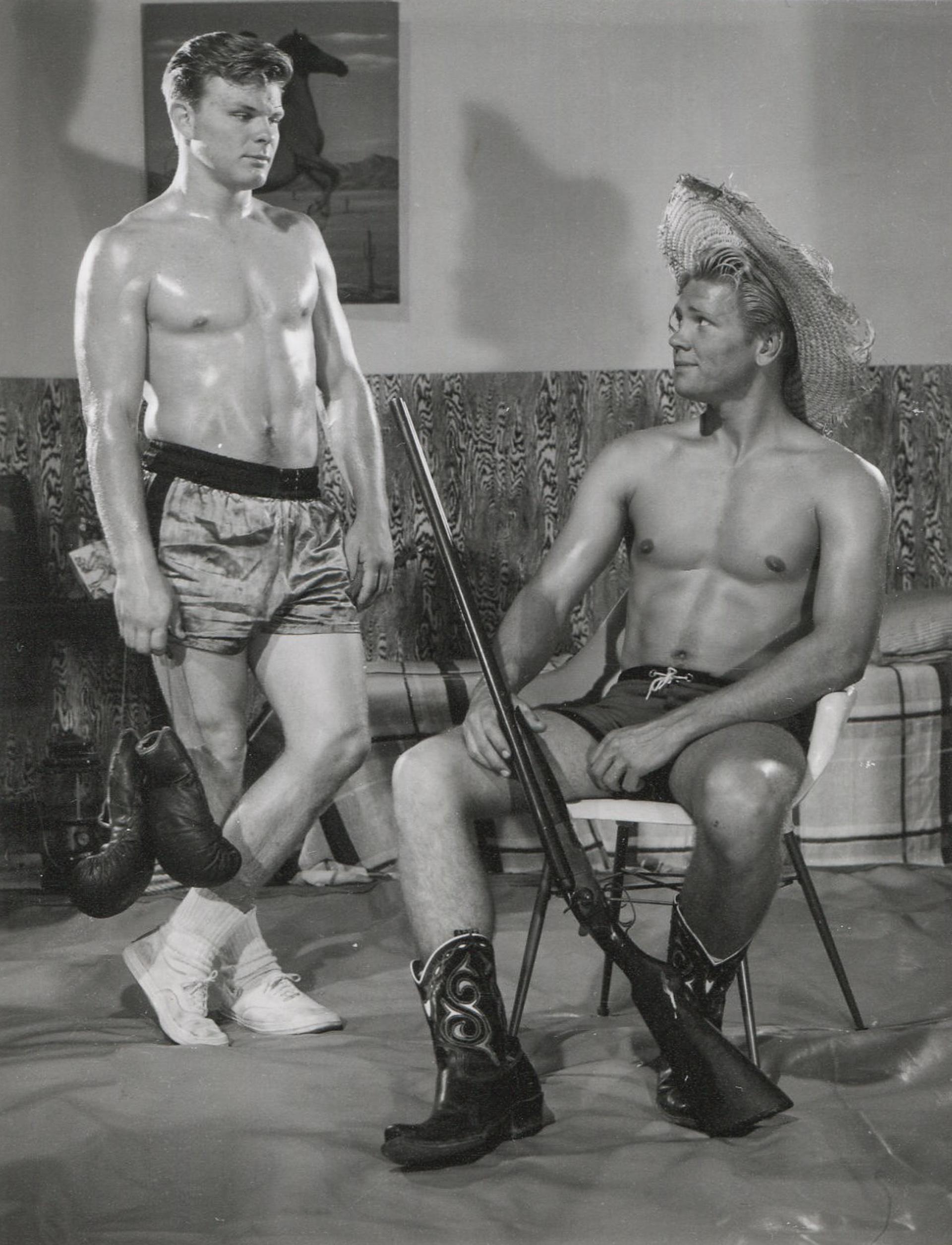 Diese Schwarz-Weiß-Fotografie von Bob Mizer, die zwei gut aussehende männliche Modelle zeigt, trägt den Titel "Ohne Titel (Zwei Männer mit Boxhandschuhen und Gewehr)". 

Untitled (Zwei Männer mit Schachtelhandschuhen und Wappen)
Verso mit Bleistift