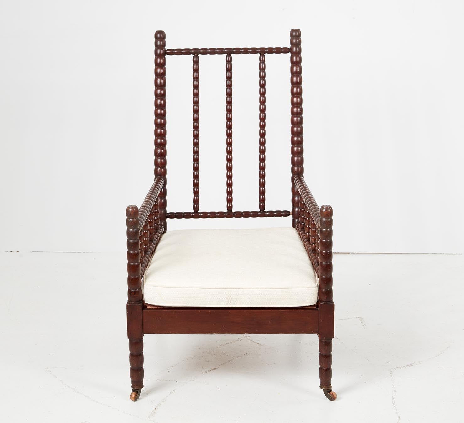 A large bobbin turned armchair with raked back on bobbin fenced arms over deep, generous seat, on front bobbin legs and shaped square section back legs with castors. Upholstered seat support. Detachable back and seat cushions in need of reupholstery