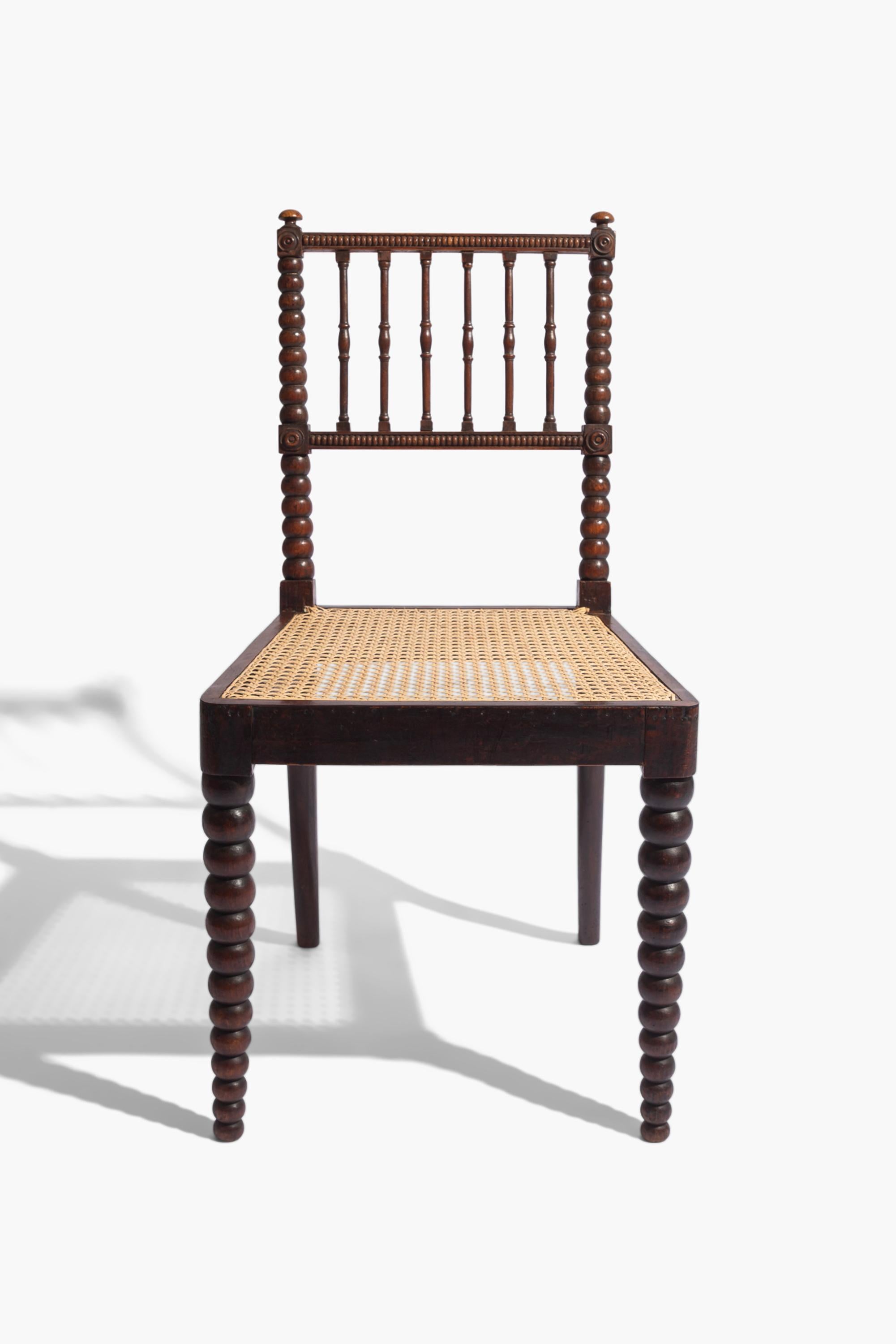 A simulated rosewood side chair designed by E.W. Godwin.

Raised on bobbin turned front legs and outswept back legs with a cane seat. The rectangular back supported by bobbin turned uprights with slender turned spindles between. Made in simulated