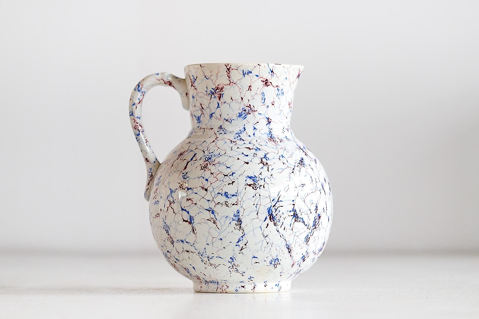 Questa brocca in ceramica smaltata è stata prodotta dal produttore belga di maioliche Boch Frères Keramis alla fine del XIX secolo. Il motivo astratto dipinto in blu e rosso è stato ispirato dai pezzi di ceramica fine prodotti in Giappone. L'effetto
