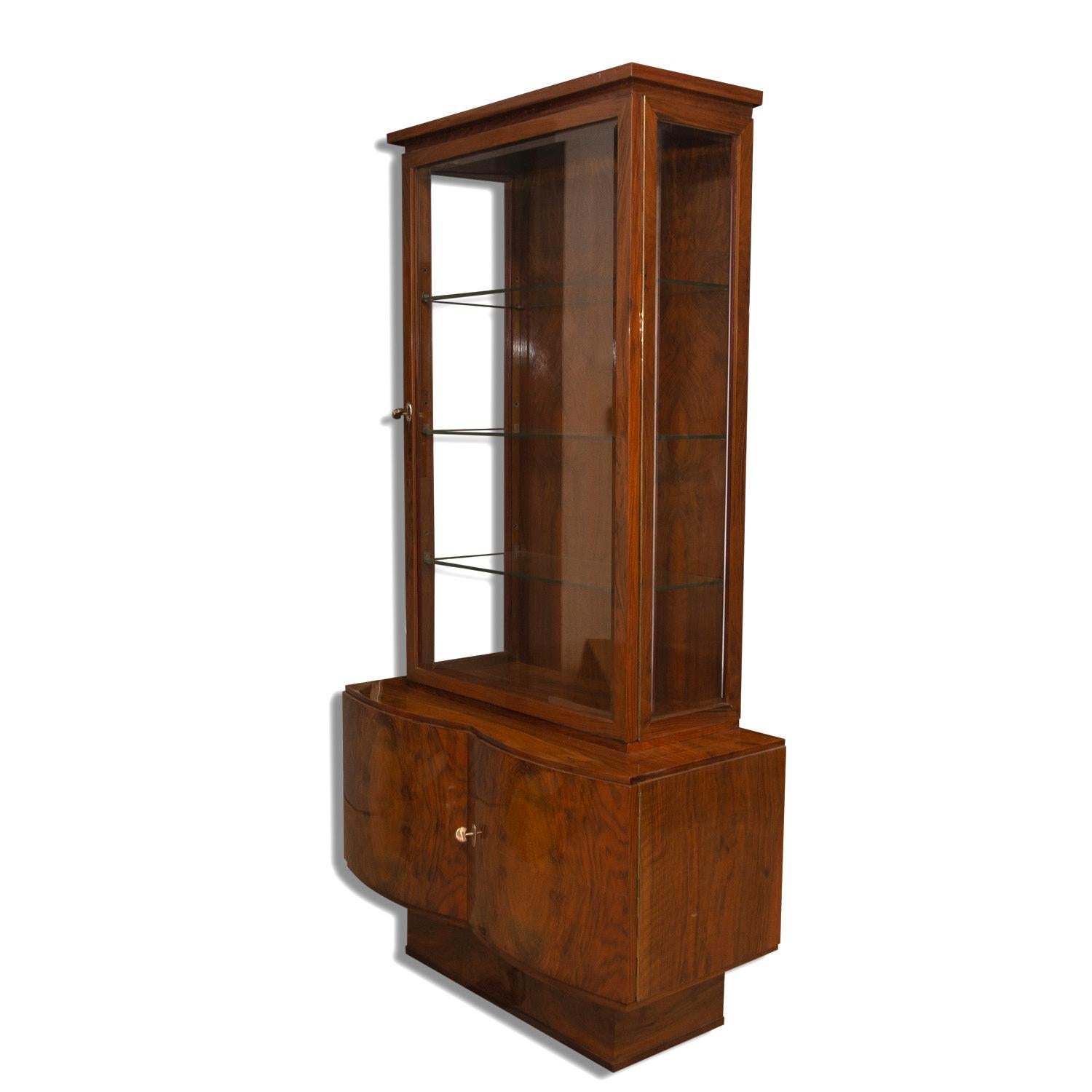 This Art Deco narrow showcase is veneered in walnut. It was designed and manufactured in the 1930s in Bohemia. The cabinet was designed in the context of the Central European pre-war design school. The cabinet has inlaid glass, rounded but also