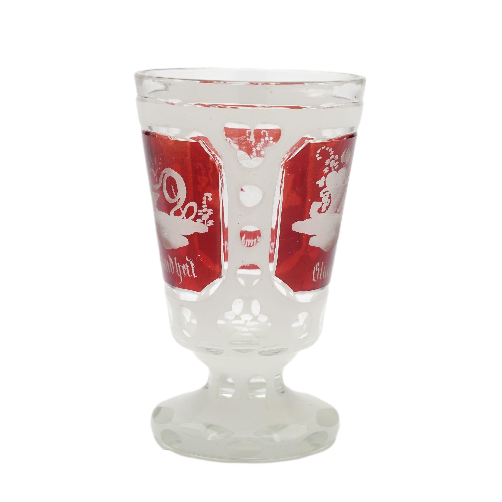 Clear blown spa glass with a cut white enamel overlay and three ruby flashed panels etched with trophies. The cup rests on a pedestal base with circular foot. Spa glasses were created for visitors to the mineral water spas to drink the spring waters