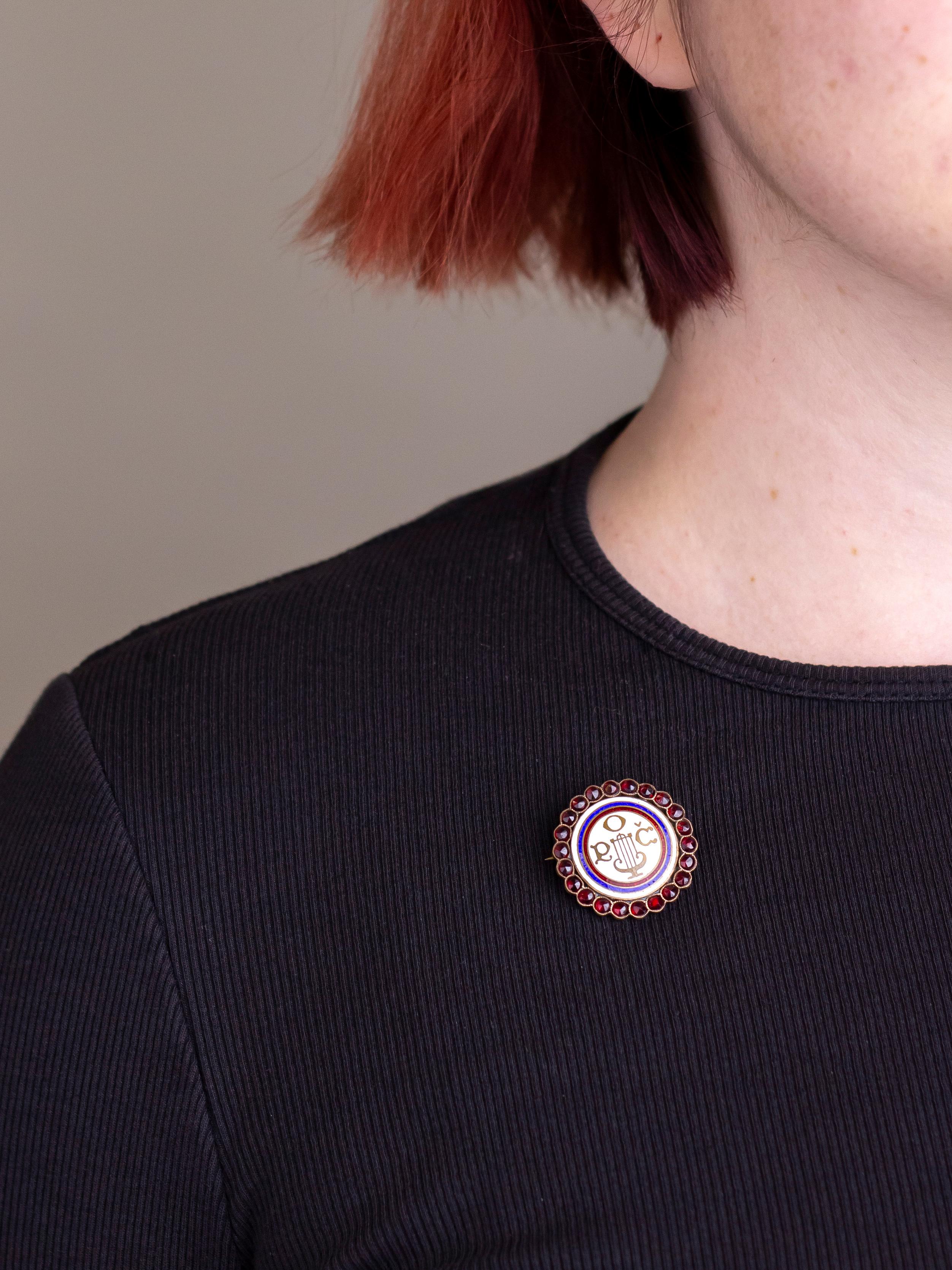 Este encantador broche engastado con esmalte y granate va acompañado de su caja original y data de principios del siglo XX. El broche redondo estilo diana se centra en las letras POC, que significan Pevecka Obec Ceska, o 