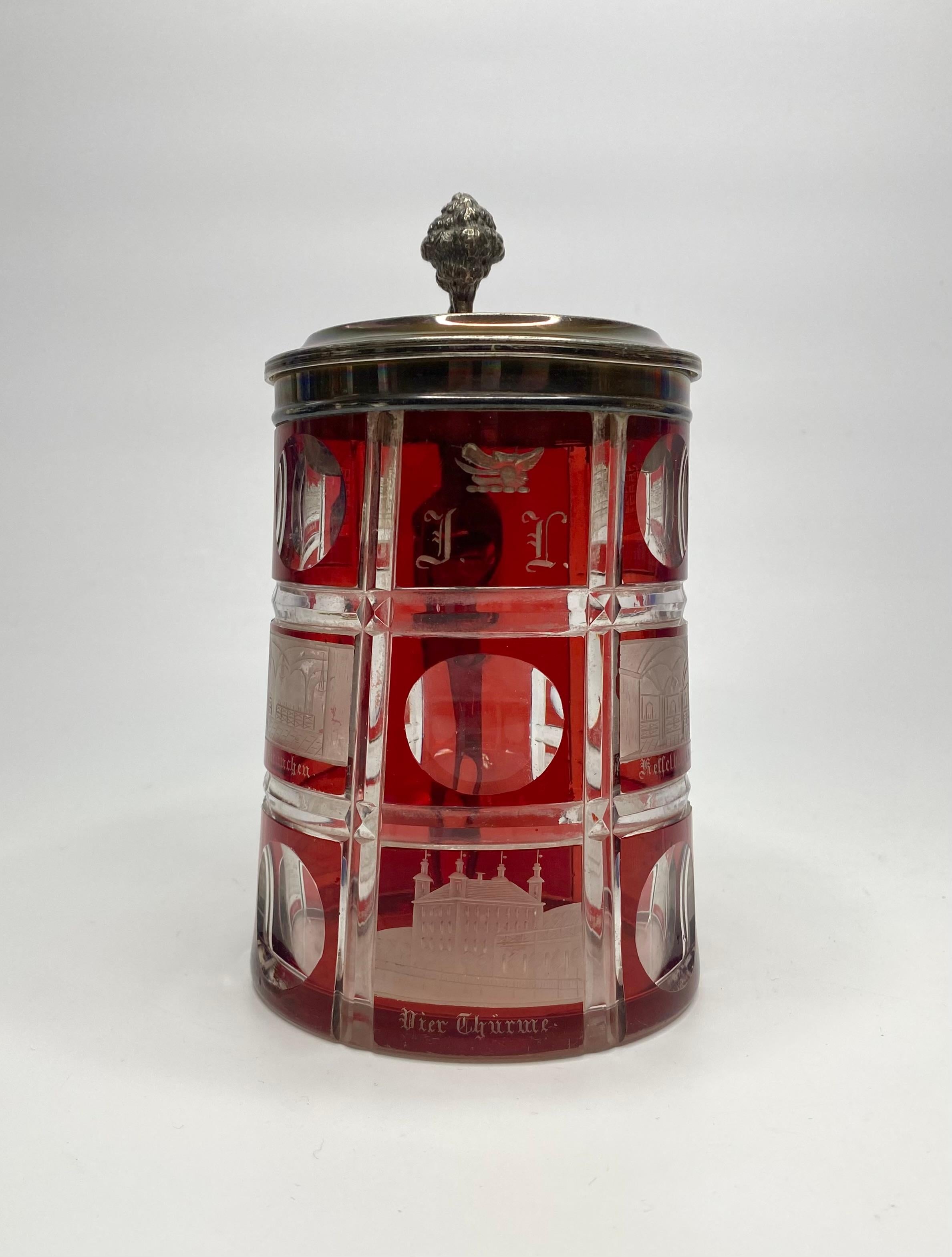 A Bohemian ruby flashed Spa glass tankard, with silver mounts, dated 1860. The large tankard engraved with panels of various buildings in German Spa towns, alternating with roundels. One panel inscribed ‘JL’ beneath a heraldic boars head. Having a