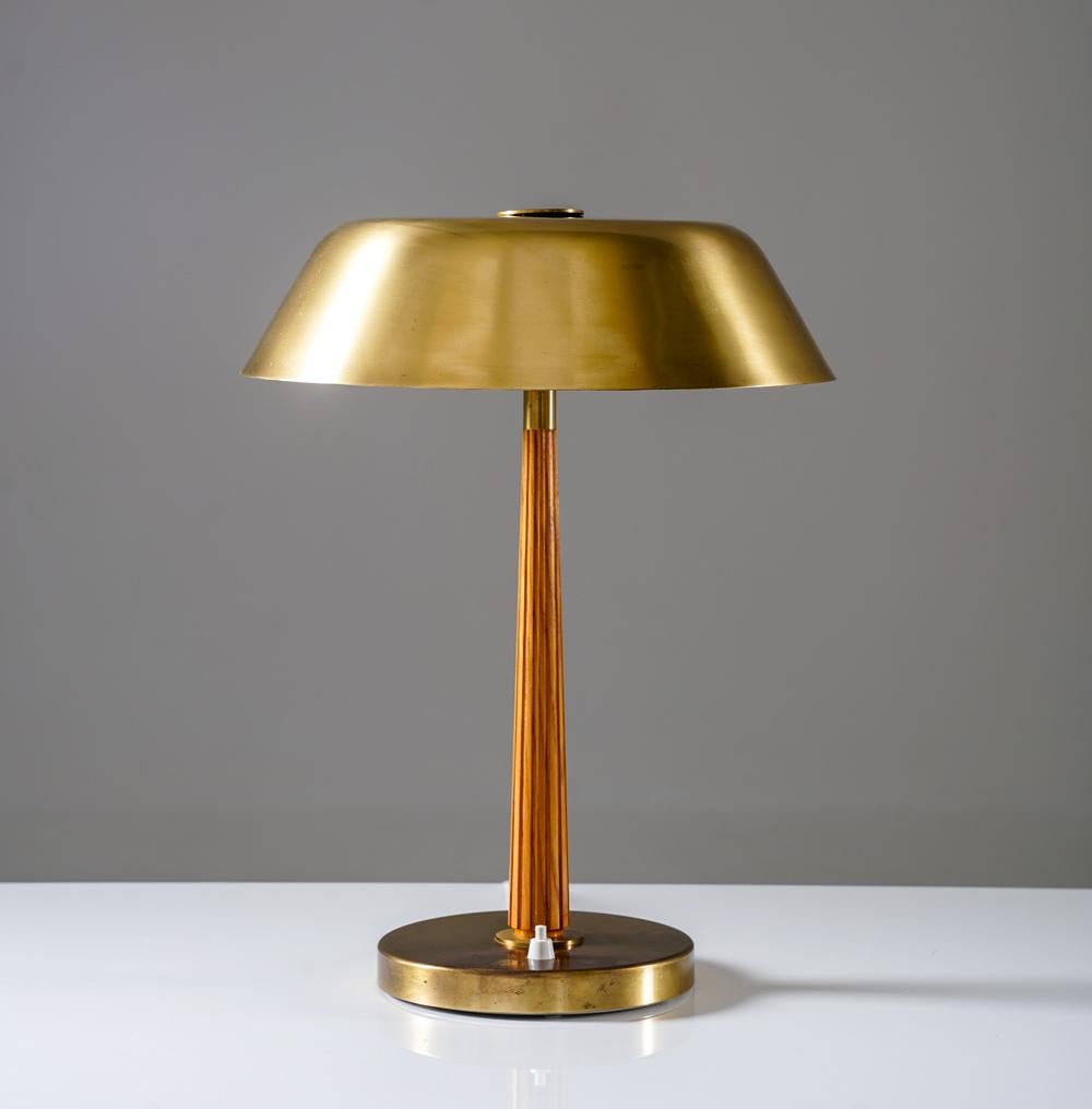 Une élégante lampe de table conçue par Harald Notini pour Böhlmarks, modèle 15474. La lampe présente un abat-jour évasé en laiton, équilibré par une tige en bois cannelée, reposant sur une base circulaire en laiton. La combinaison de laiton chaud et