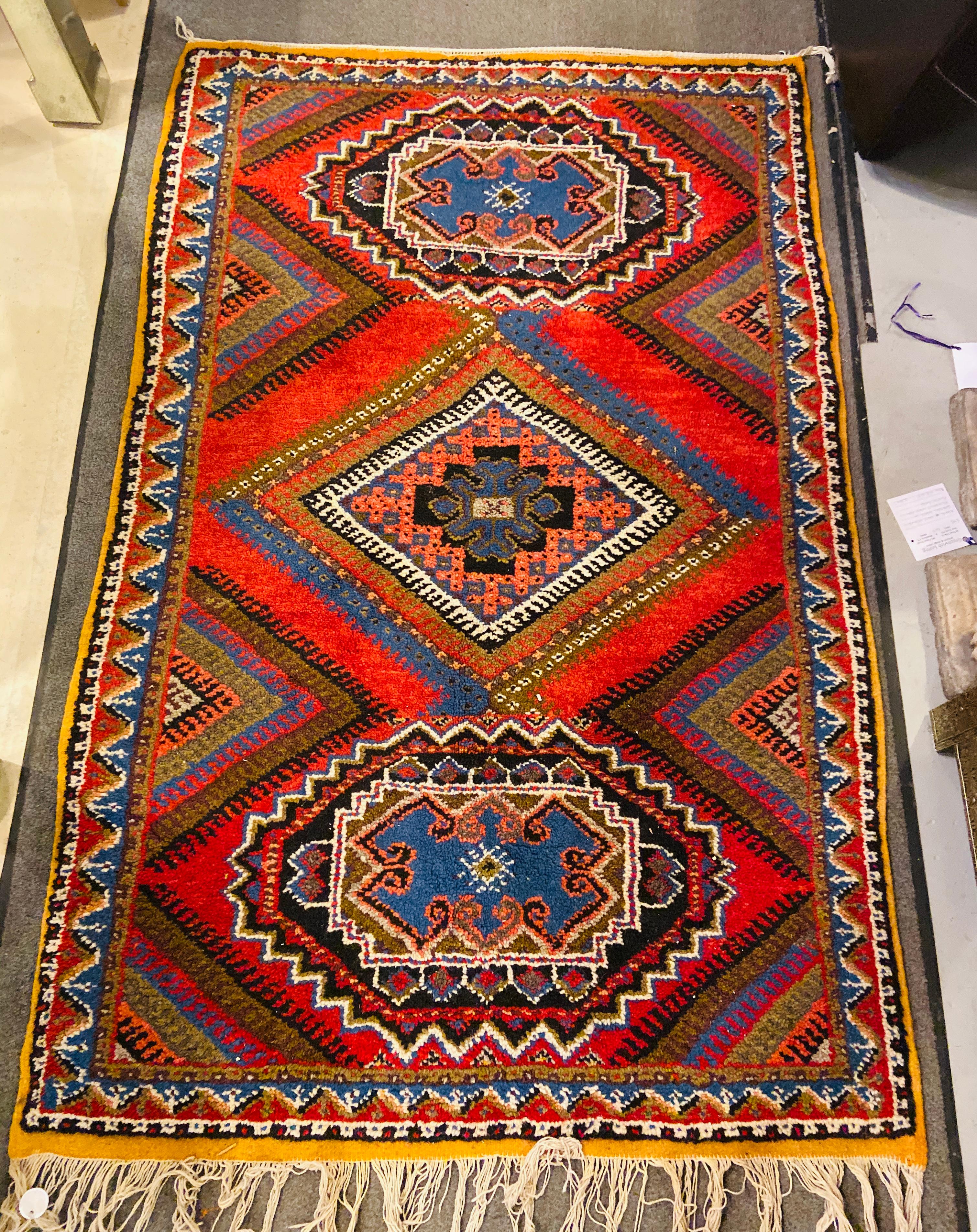 This is a vibrant Moroccan handmade rug featuring a rich and intricate design typical of traditional Berber craftsmanship. The rug has a rectangular shape and showcases a dominant color palette of reds, blues, dark greens, and whites.  At the