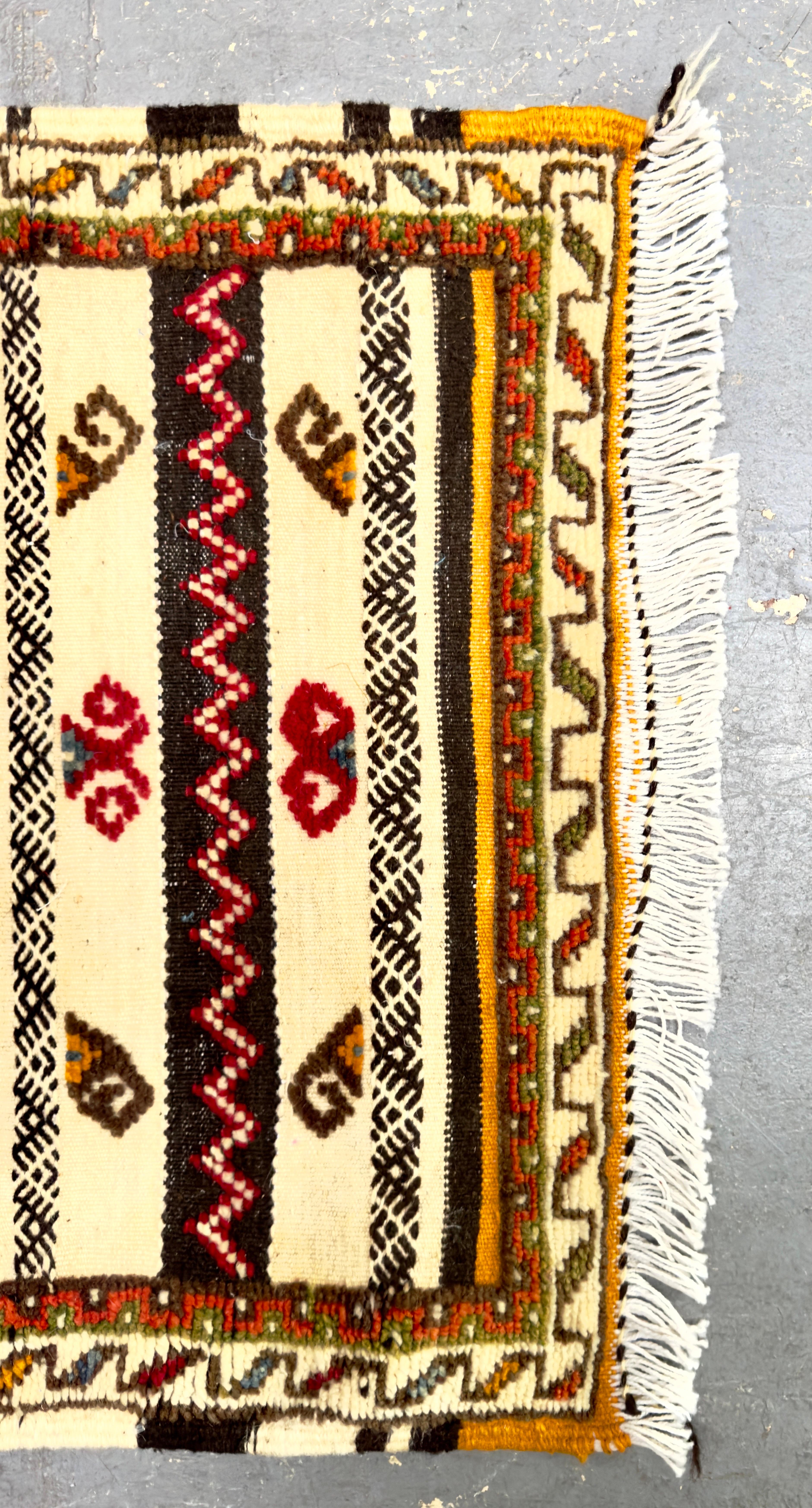 Der marokkanische Teppich Boho Chic zeichnet sich durch ein traditionelles Design mit komplizierten Mustern und einer lebendigen Farbpalette aus. Das zentrale Feld ist ein markantes rotes Rechteck mit einem geometrischen Rautenmotiv in der Mitte,