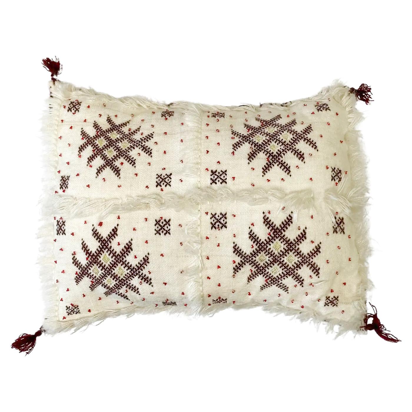 A stunning pair of boho chic Moroccan Handwoven pillows boasting magnificent Berber geometric patterns and refined fringe work. The white pillows are made of soft wool and embellished with red beads and dark purple berber geometrical motifs. The