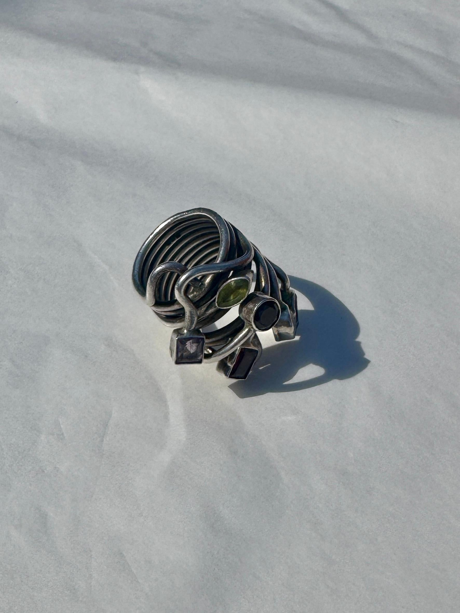 This striking boho ring in 925 sterling silver features a sculptural, wrap-around design made of multiple hammered bands that gracefully coil around the finger. At the top, flowing, organic silver branches hold a selection of natural gemstones in