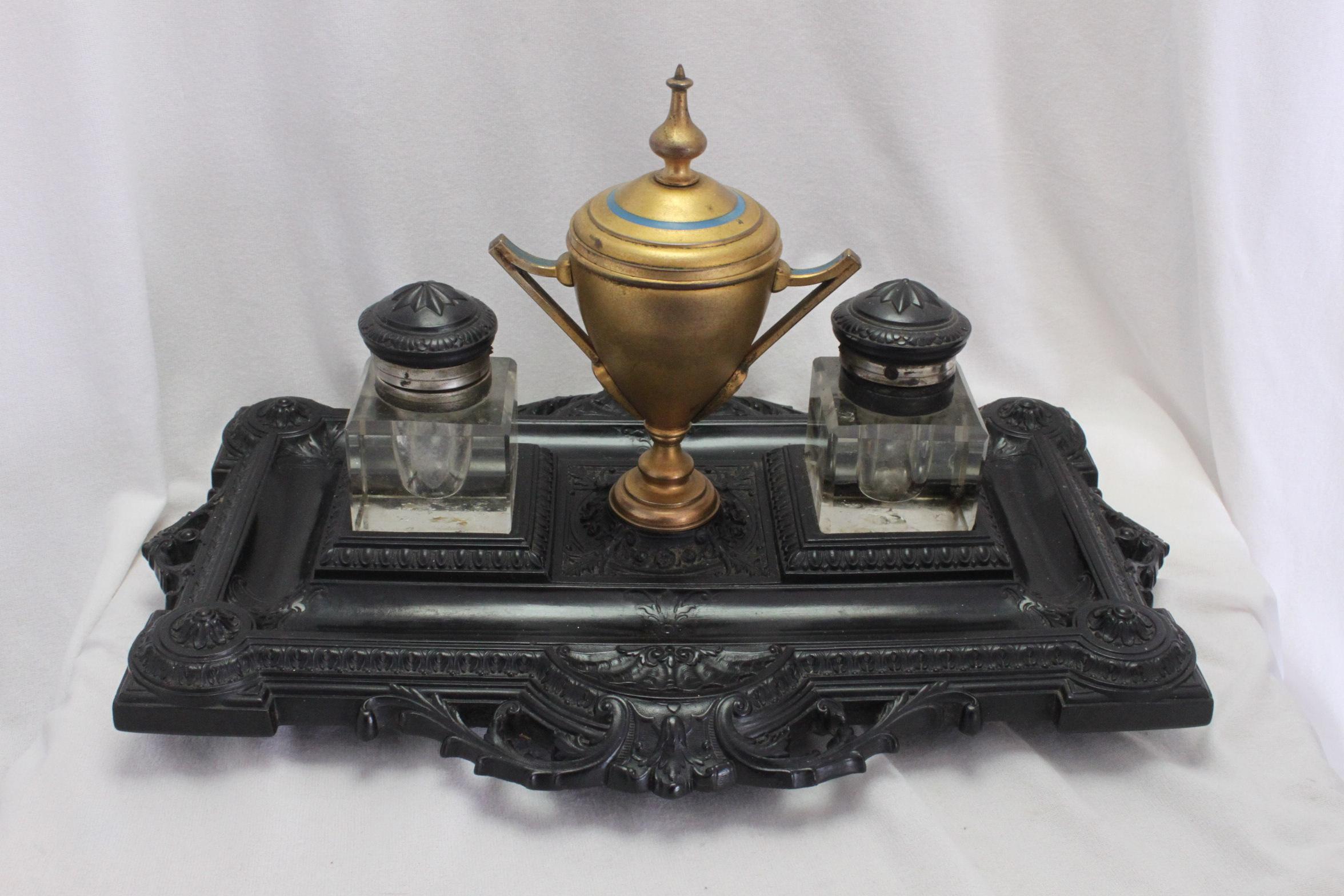 This large Bois Durci ink stand features an intricately moulded base, upon which sit two glass inkwells and a central urn shaped container. The glass inkwells are fitted with bois durci tops set into silver plated mounts and collars. The urn shaped