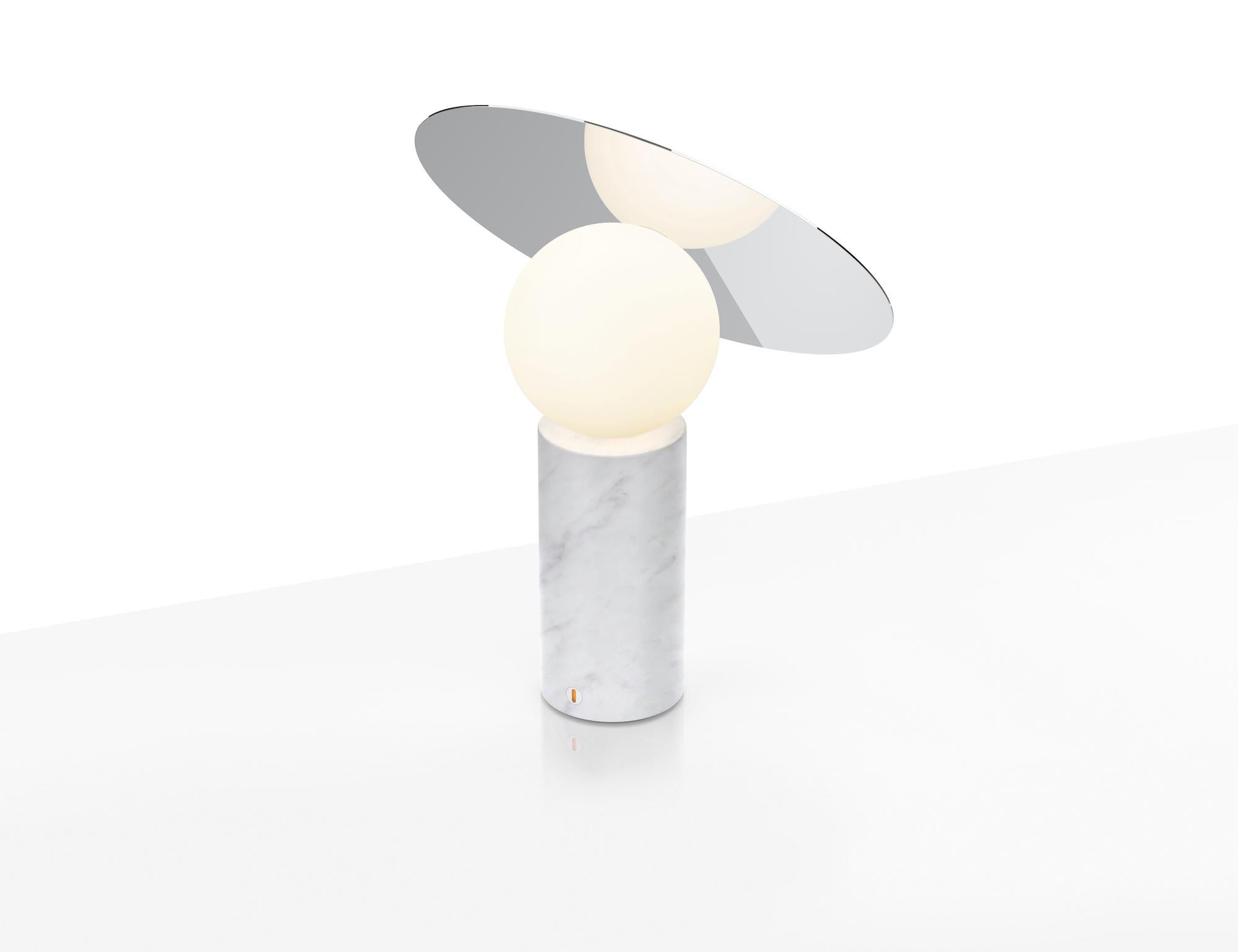 La icónica silueta de la mesa de disco Bola está definida por su elegante pantalla de disco de acero inoxidable cromado colocada a 45 grados y elevada por un difusor de globo de cristal opalino que parece flotar sobre su base cilíndrica de mármol.