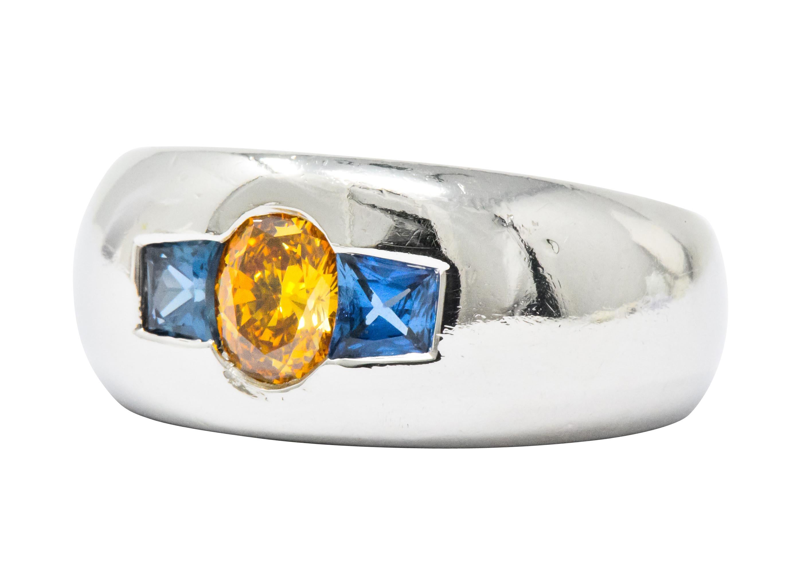 A wide, flush set three stone band ring with a domed curvature and bright finish

Centering an oval cut fancy colored diamond weighing 0.59 carat, intense yellow-orange in color with VS1 clarity

Flanked flushly by two calibre cut sapphires weighing