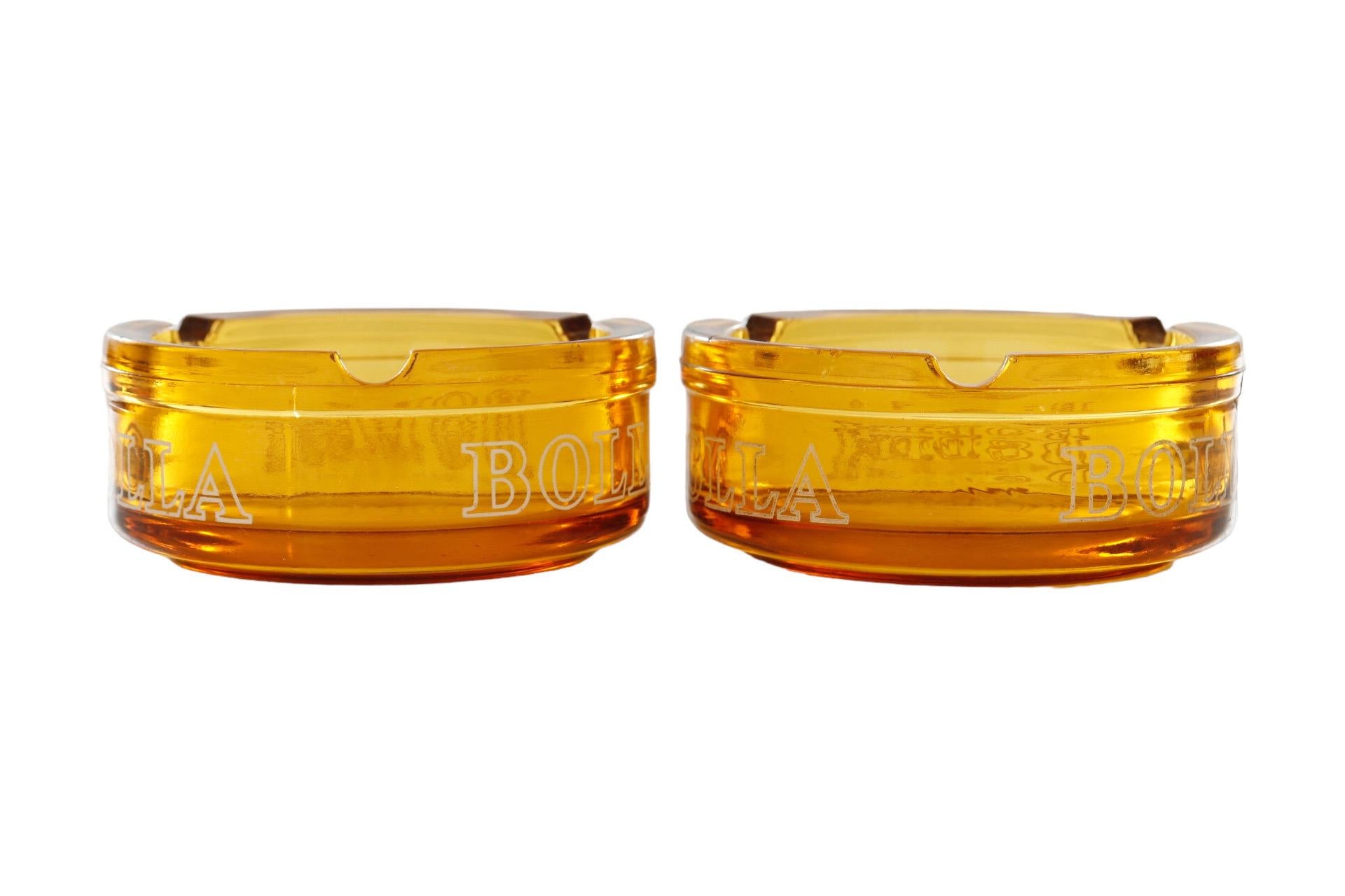 A pair of Bolla wine amber glass ashtrays. Each ashtray has three cigarette rests and 'Bolla' in bold white lettering repeated three times around the edge. Dimensions per ashtray.
