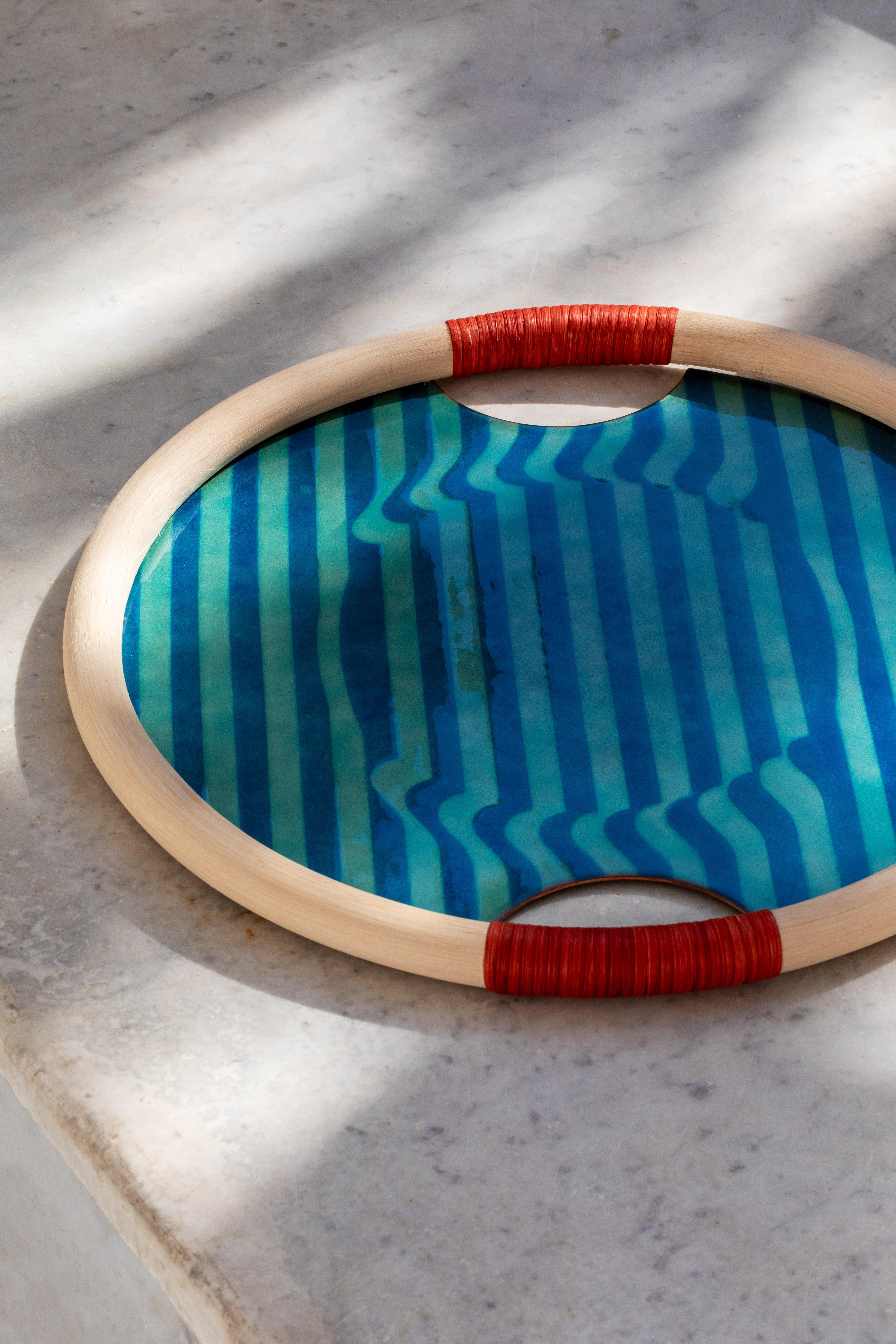 Bombé is a round enameled copper tray with an optical design that recalls the waves and colors of the lagoon, set in a frame of manila, the pith of rattan. The handles are embellished with two bombé rattan weaves, in red Incalmi.