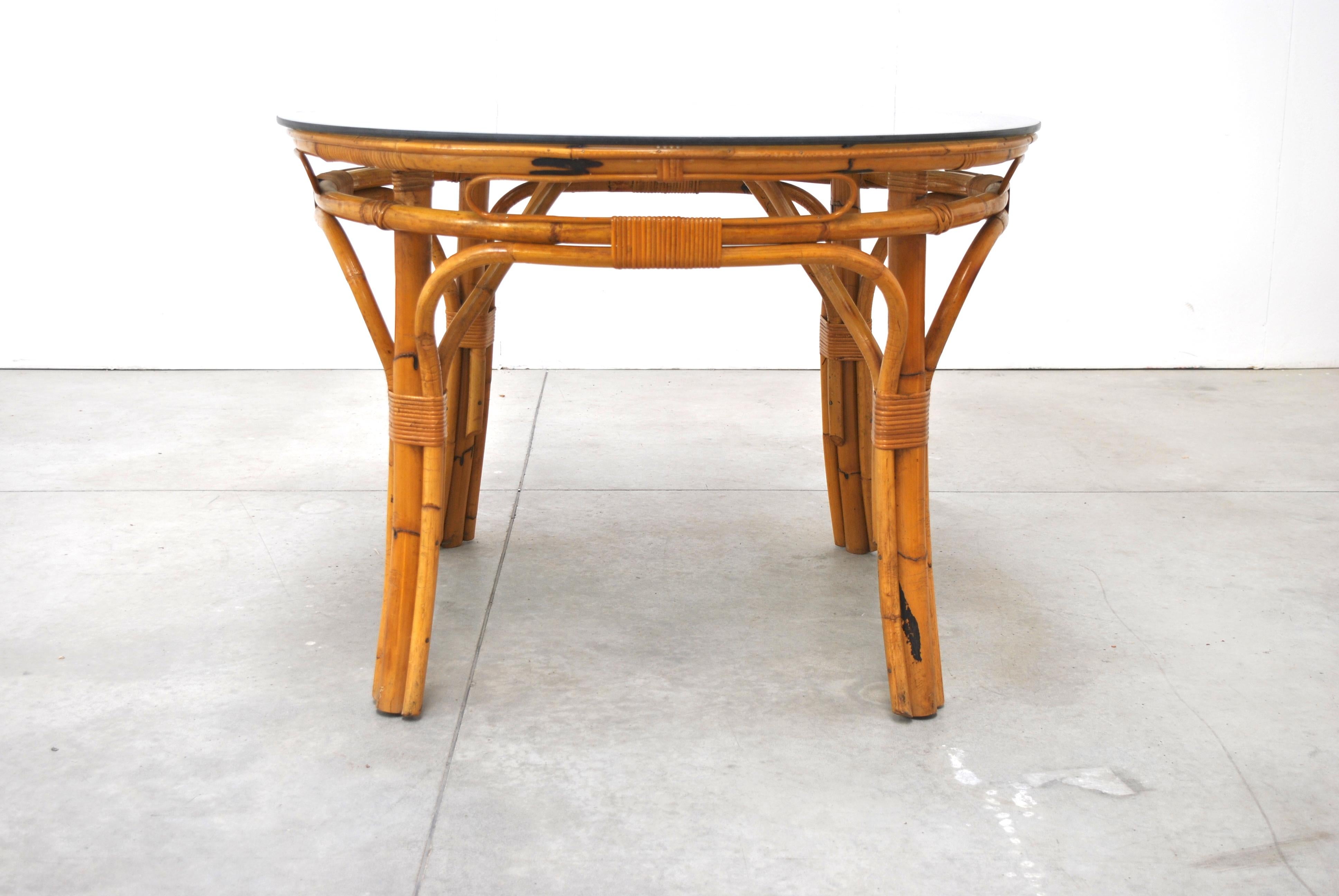 Mesa de comedor de ratán de mediados de siglo. Tiene un gran tablero redondo de cristal y la mesa se apoya en patas redondas de bambú reforzadas con flejes de bambú.