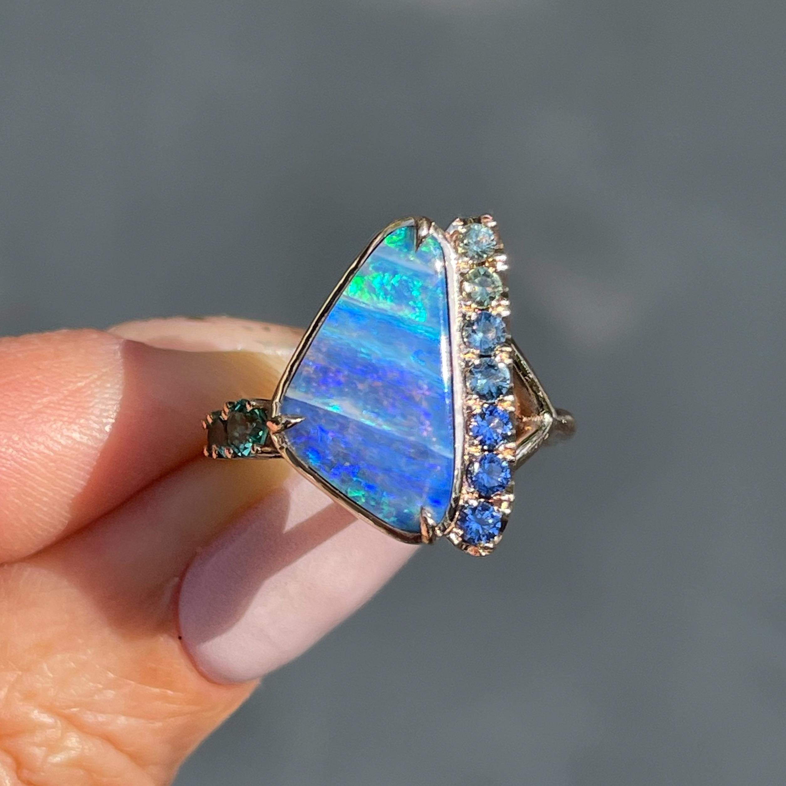 Ocean blues and seafoam greens traverse lanes in this Australian Opal Ring. Pooled hues in the opal stone are flanked by plunging ombré sapphires and a surge of emeralds in vivid green. The Boulder Opal Ring imposes a sense of order upon nature. Yet