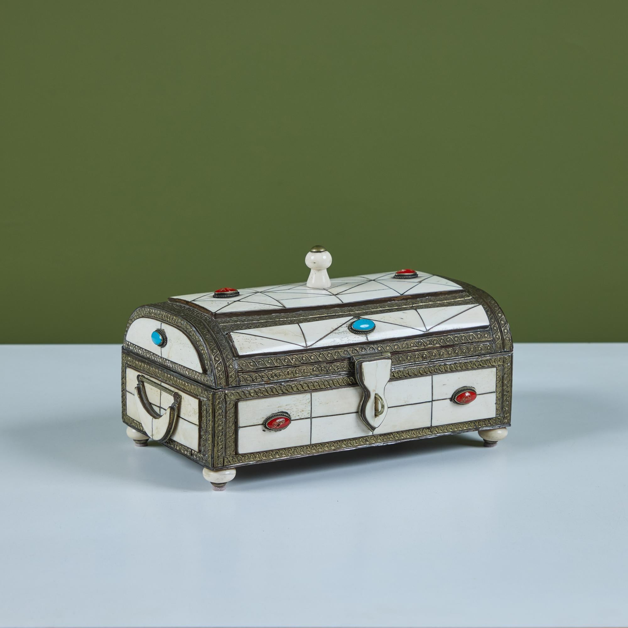 Caja decorativa de metal con incrustaciones de hueso y piedra. El exterior de la caja muestra una incrustación de hueso en mosaico adornada con turquesas y piedras rojas engastadas en peltre. El interior de la caja está forrado de cuero marrón. Cada