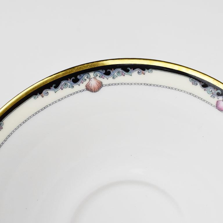 A beautiful crisp white bone china saucer with a sea shell design around the rim. It has a crisp white center, and a hand painted gold rim. Light pink shell designs are painted atop a cream background around the edges. Ready to mix and match with