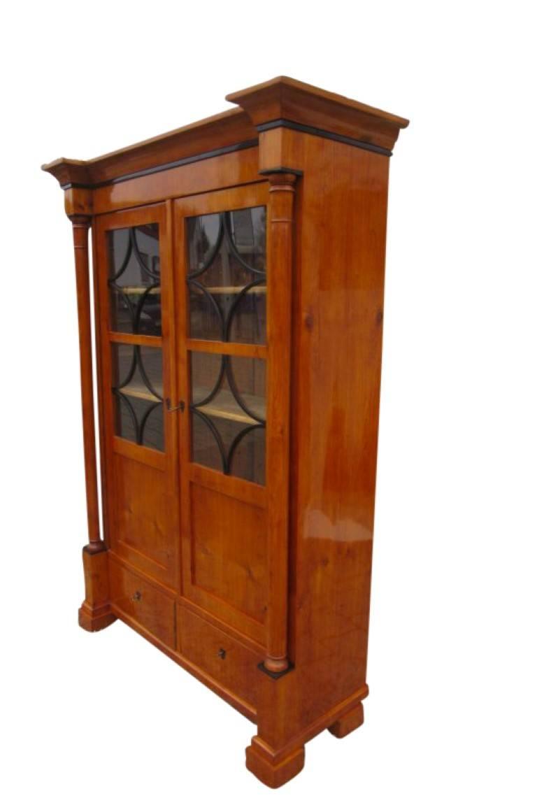 A bookcase in Biedermeier style from 1900. The corpus is made of softwood with a cherrywood veneer. The furniture is healthy and without worm damages despite the age. In the base of the piece are two drawers and above that two doors with glass