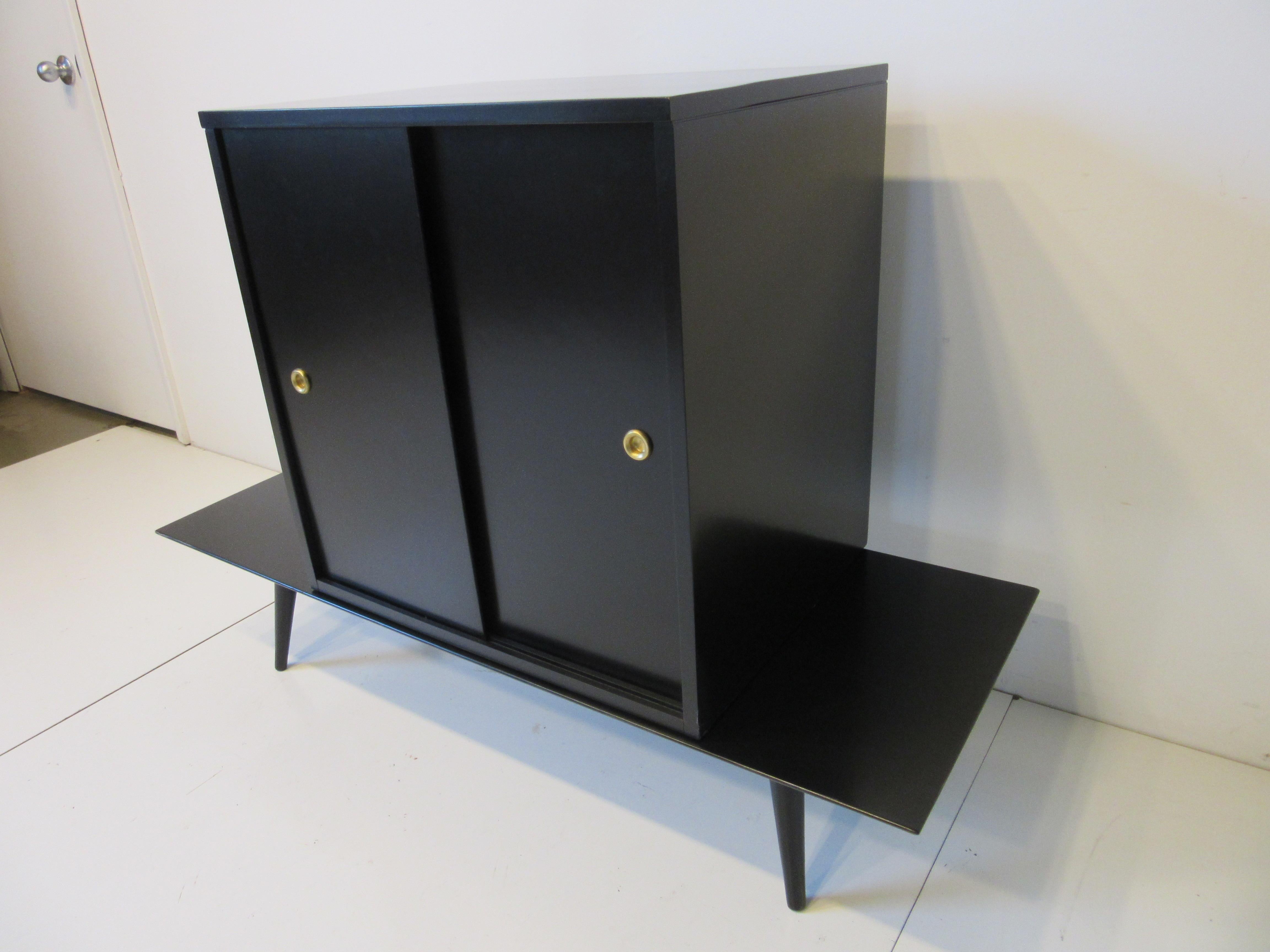 A cabinet sitting on a bench base with conical legs, having one fixed shelve with sliding doors and storage finished in satin black having brass finger pulls. Solid maple wood construction manufactured by the Winchendon Furniture company from the
