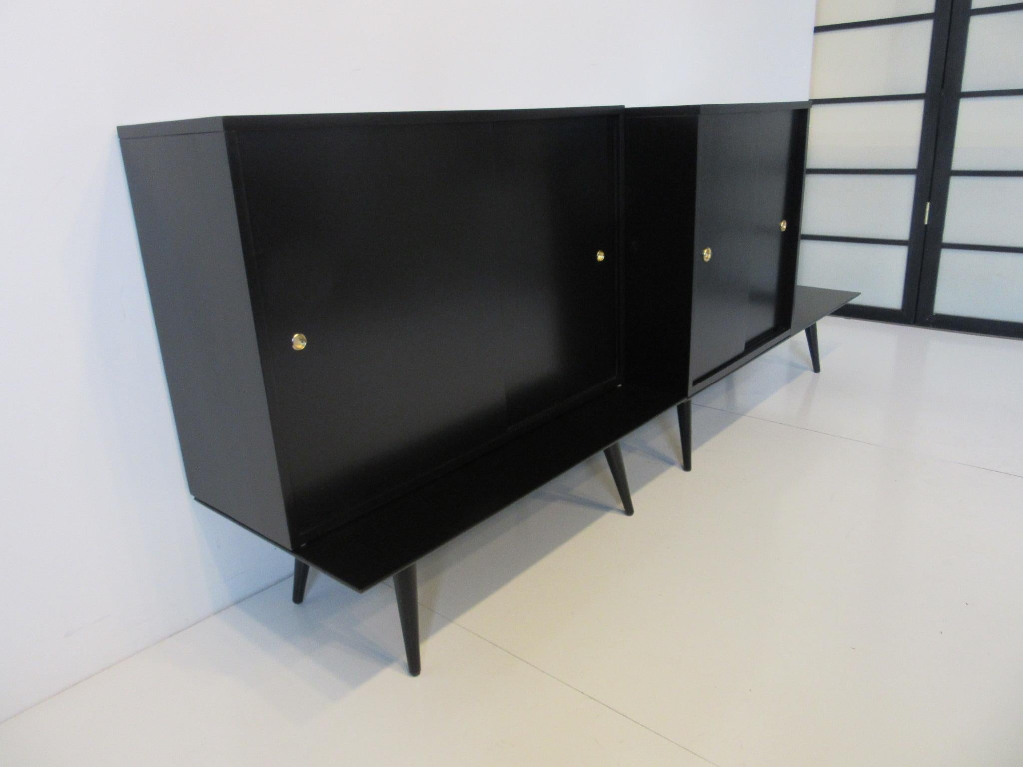 A four piece cabinet set sitting on two bench bases with conical legs, having one bookcase with sliding doors and a storage chest cabinet also with sliding doors finished in satin black and having brass finger pulls . In solid wood construction