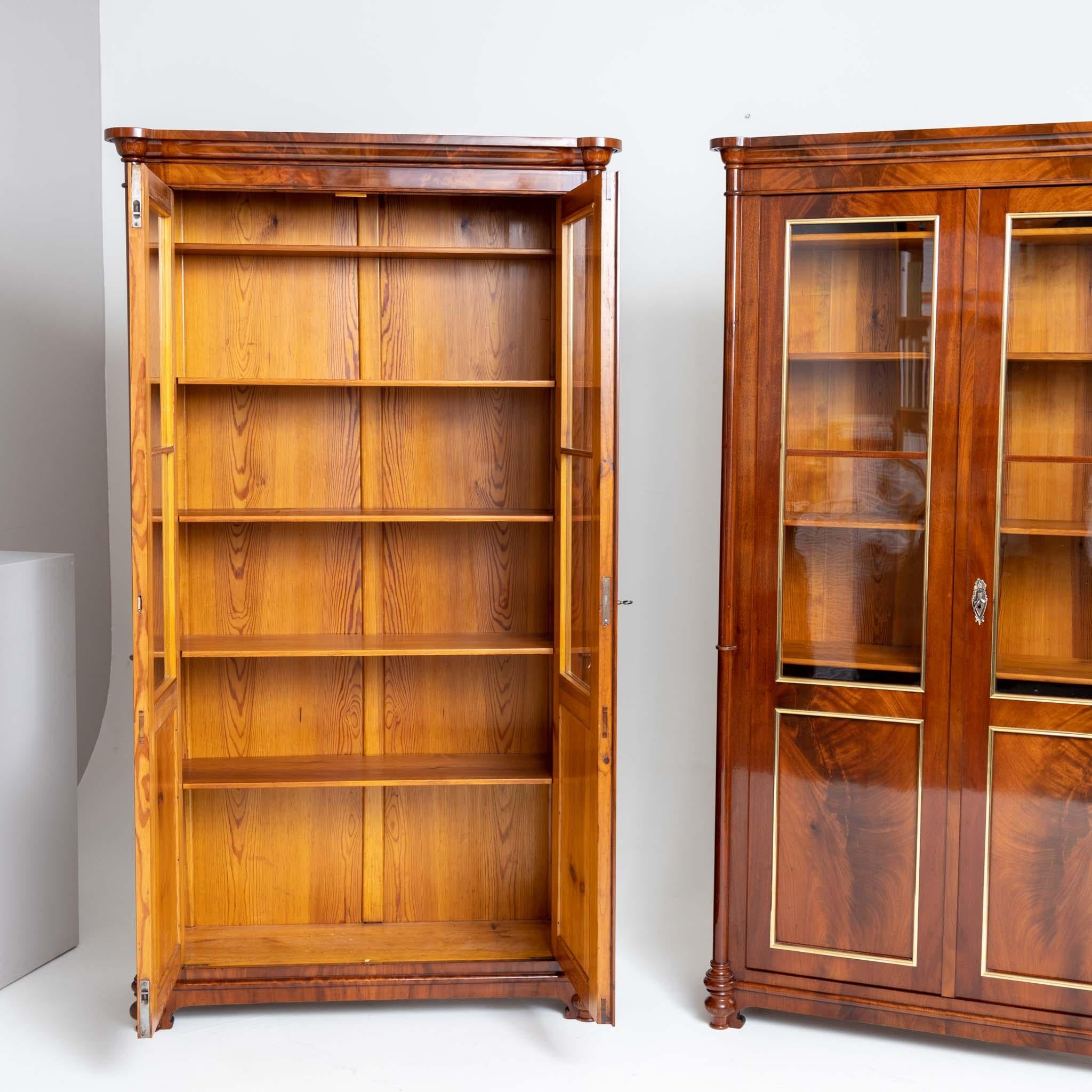 Two mahogany veneered bookcases with glazed doors and brass moldings, executed as a close pair. The corners are designed as three-quarter columns with central ring and profiled bases and capitals. Internal dimension (d): 24cm. Restored and
