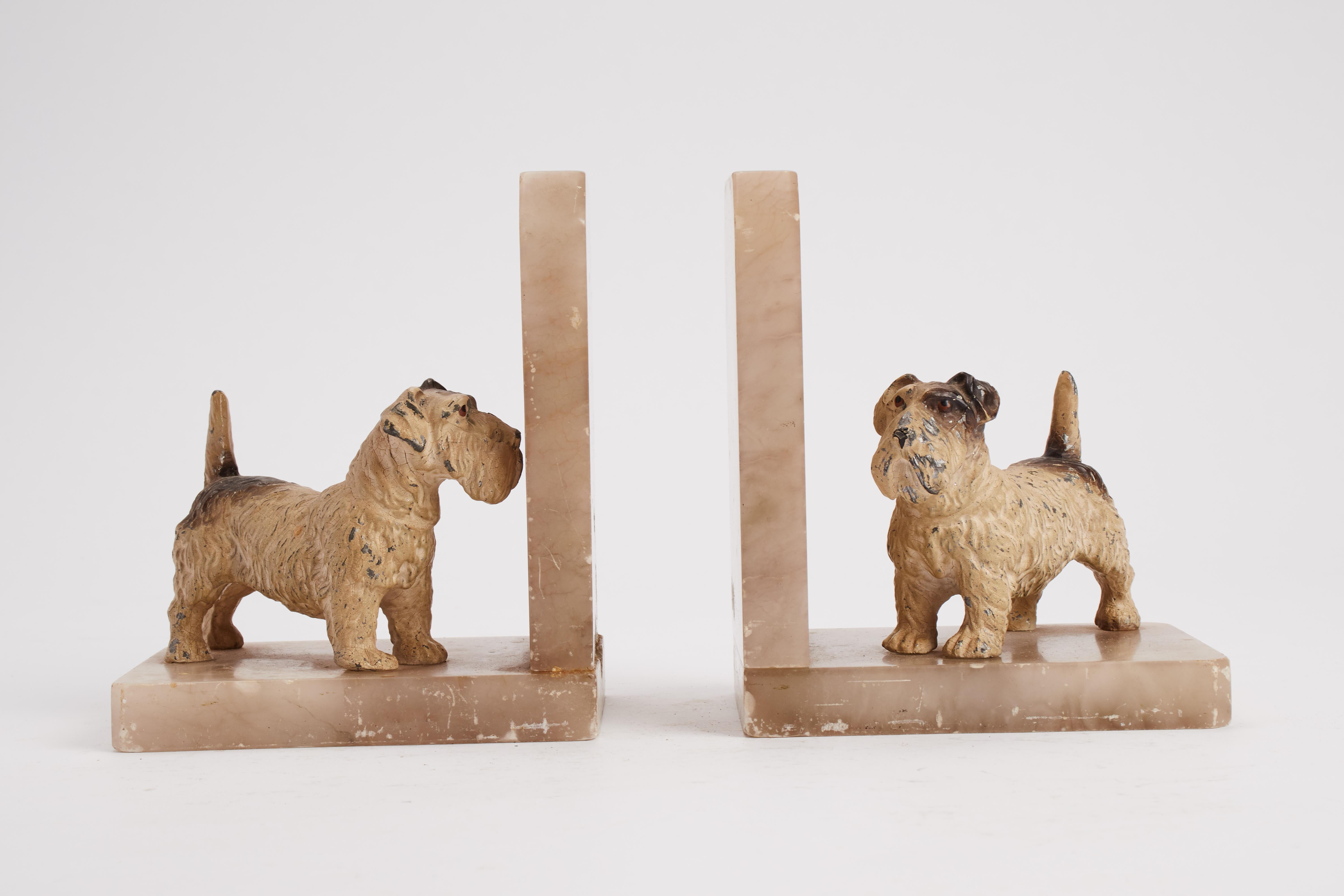 The pair is identical, showing a pale marble base with two elements at 90 degrees. Over the base, two white metal sculptures, color finished, depicting Sealyham terrier dog. England, 1920.