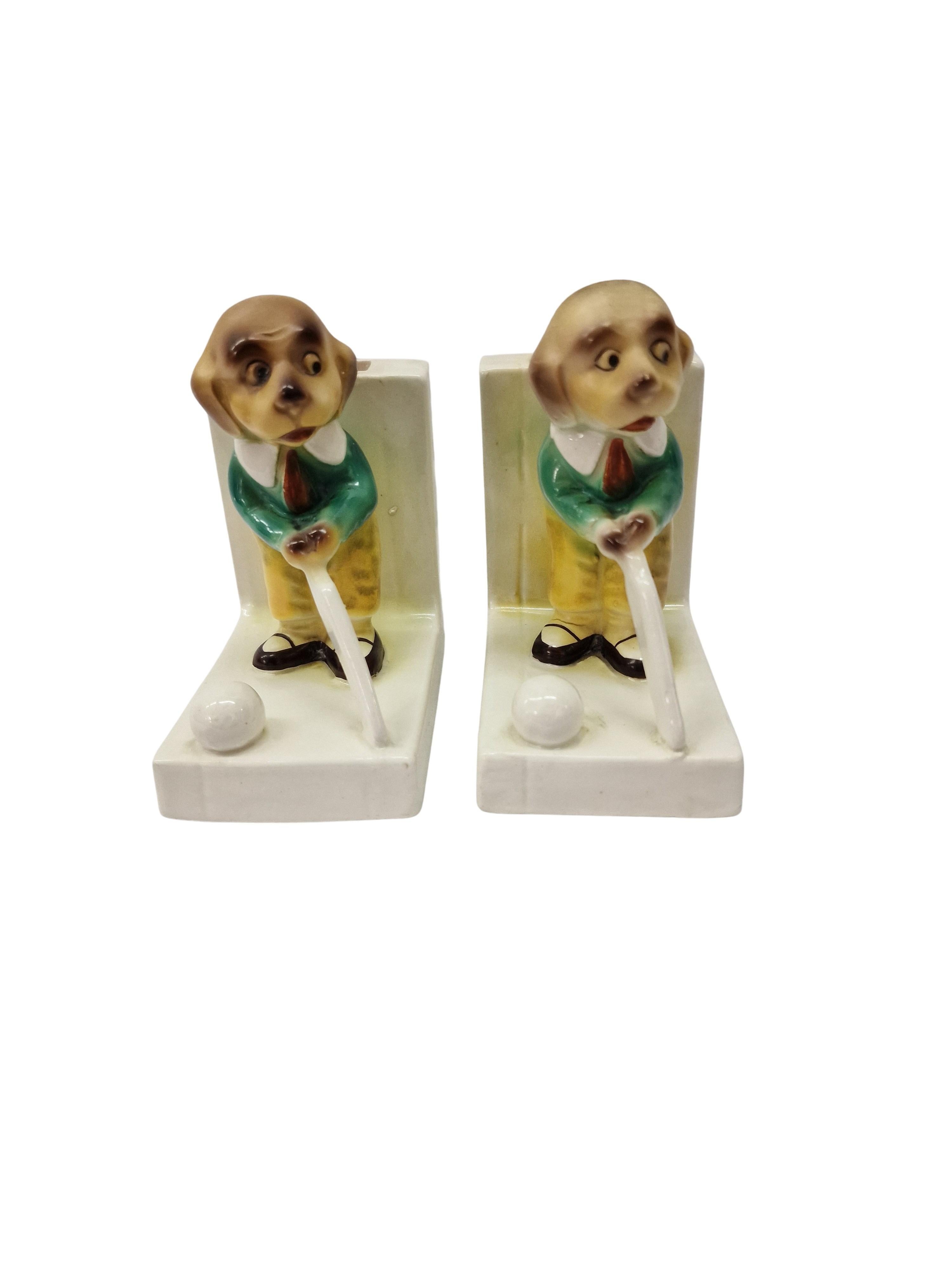 Exceptionally charming pair of porcelain bookends made in England before 1923. 

The bookends are L-shaped and the high side also has an opening as a vase for flowers or dried flowers.  Each stand features a dog in traditional clothing, a green