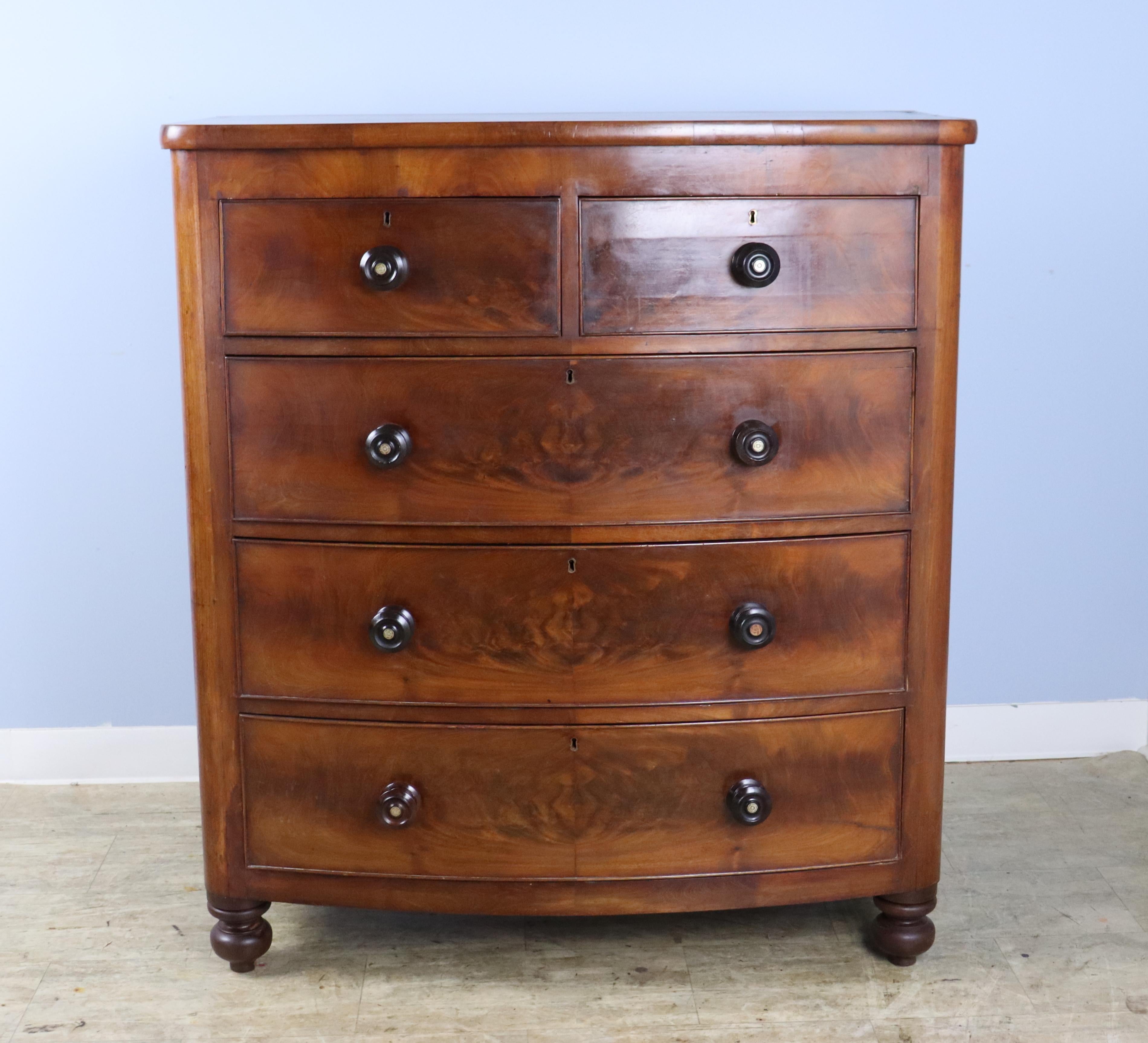 A handsome bowfront chest of drawers with bookmatched flame mahogany veneer.  Other features of note are the eye catching knobs, which may be replaced, the sweet bun feet, and the original blue paper drawer lining.  There is some buckling of the